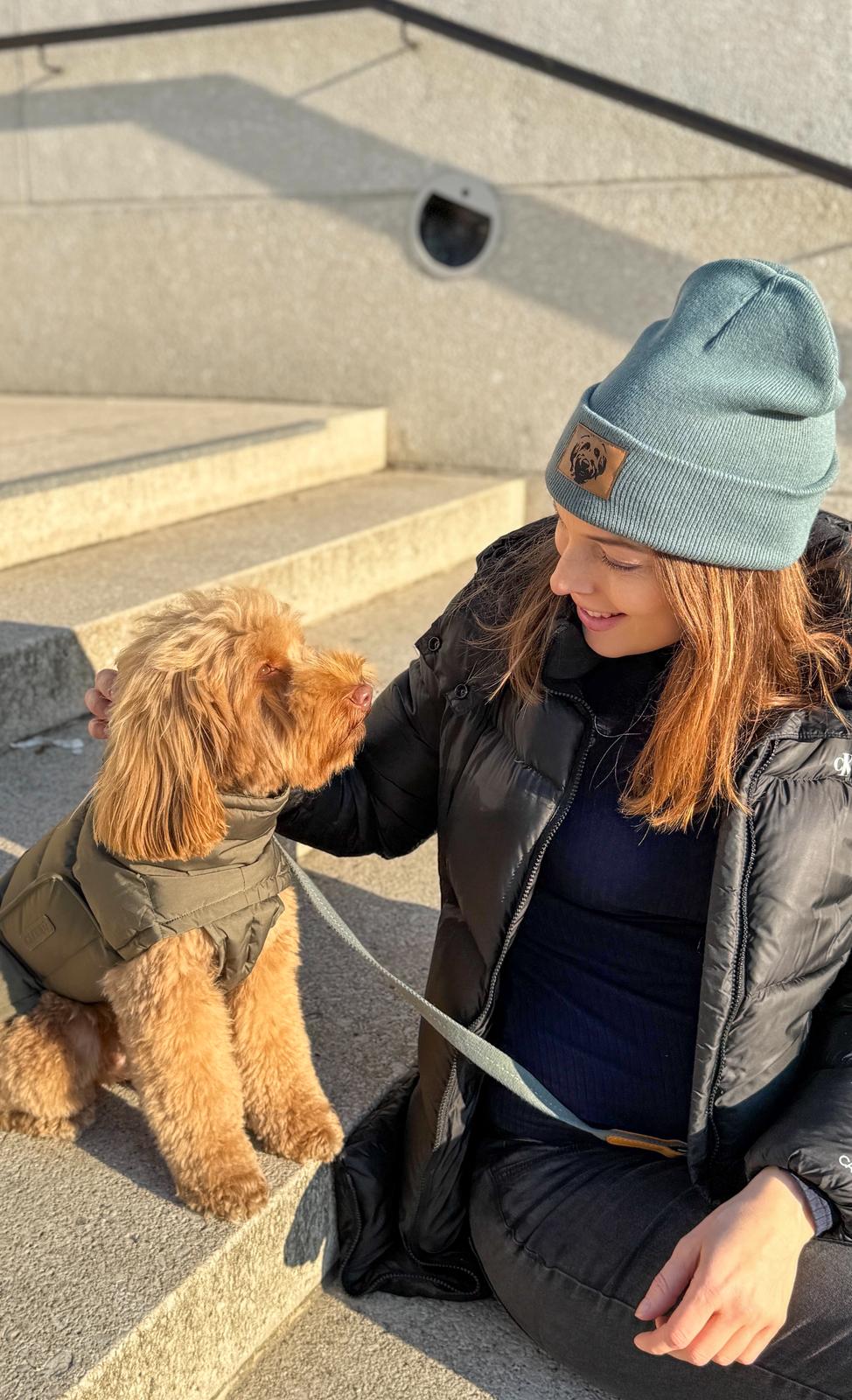 Frau mit salbeigrüner Strickmütze mit Labradoodle-Logo streichelt ihren Hund im Freien – gemütliche Beanie für Spaziergänge mit Hund bei kühlem Wetter.
