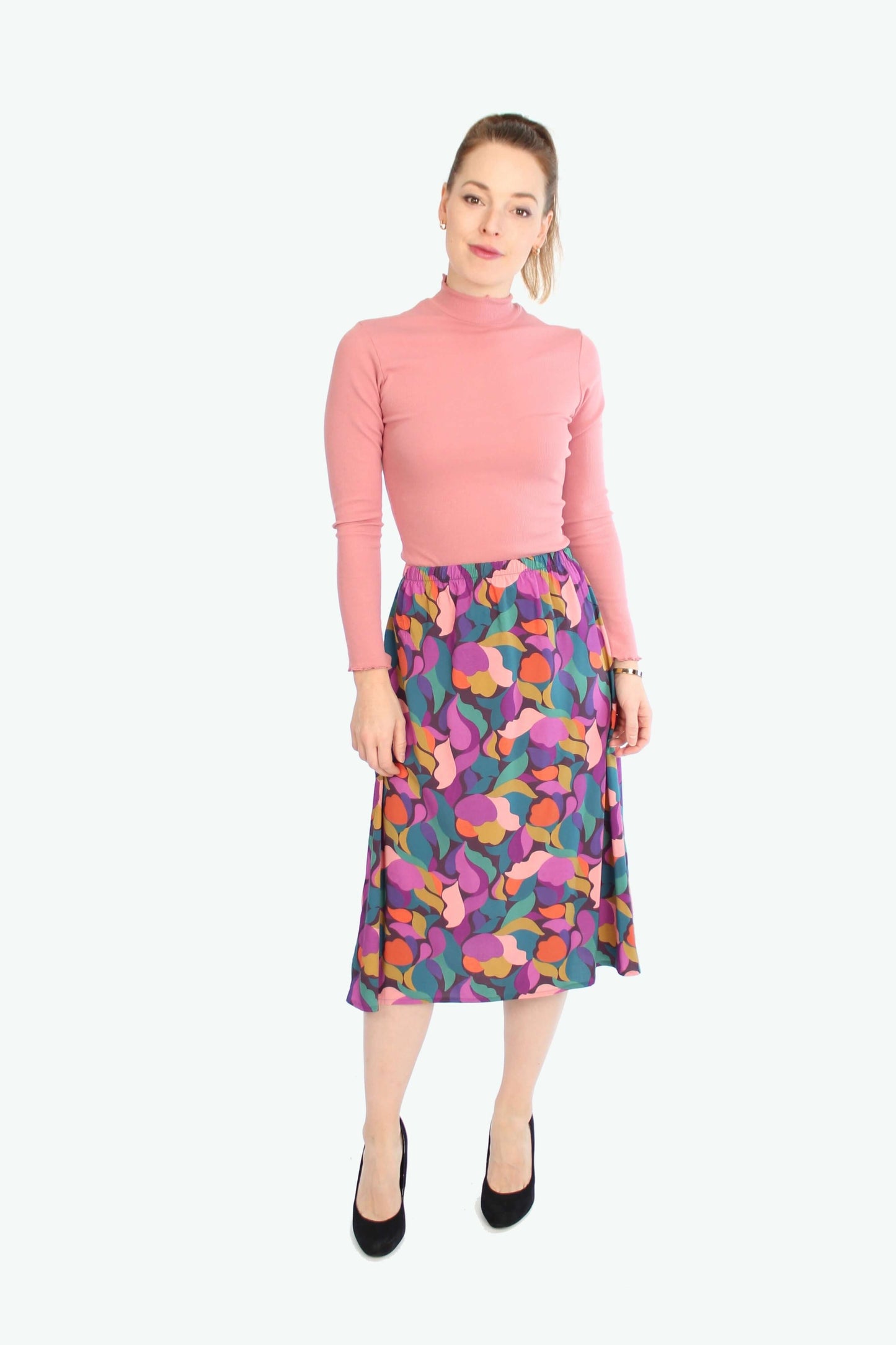 LA.LUKA Berlin Midirock mit floral-abstraktem Muster in leuchtenden Farben – kombiniert mit rosa Langarmshirt und schwarzen Pumps.