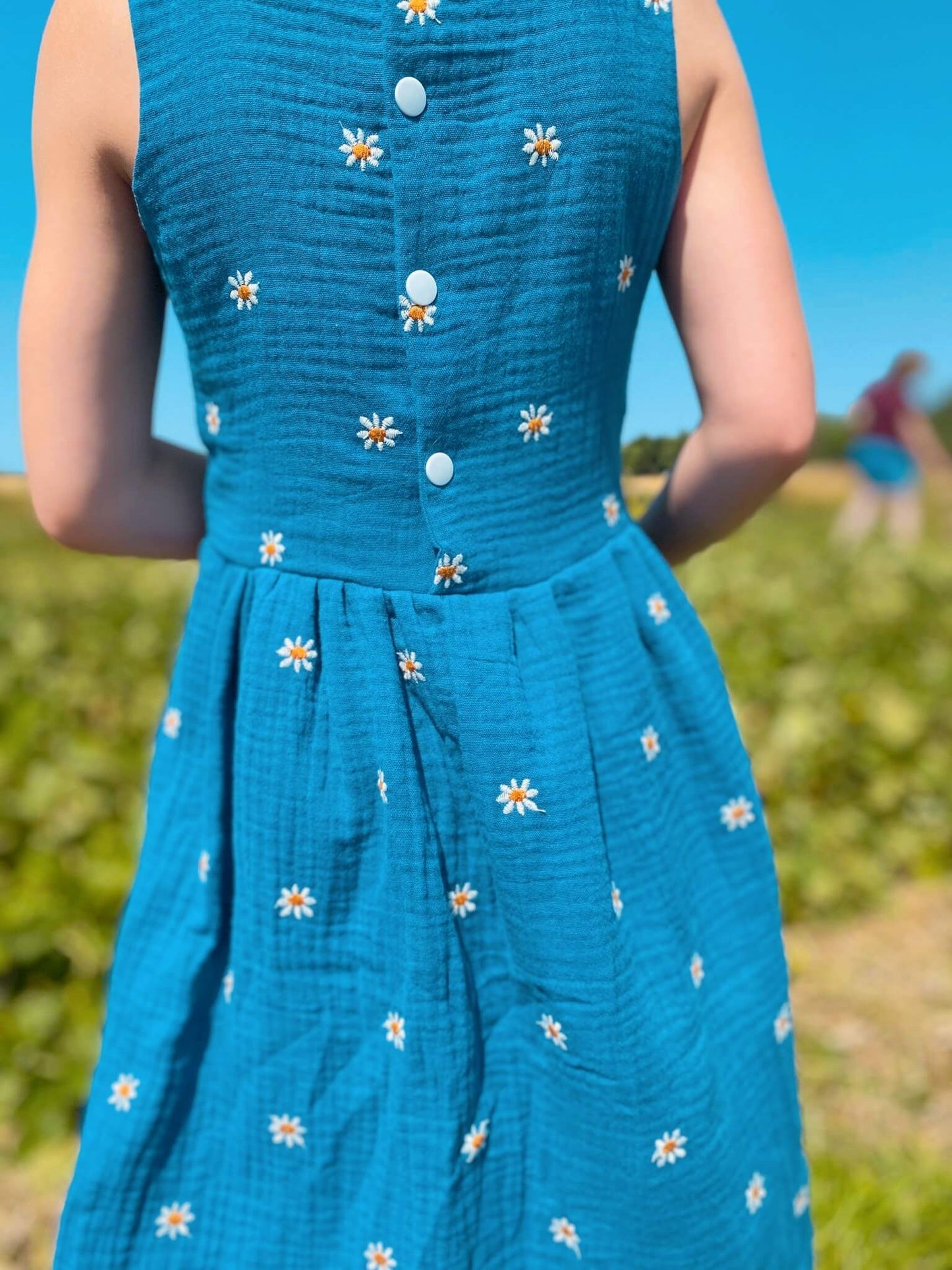 Mädchen in einem Kleid in blau mit weißen Gänseblümchen aus Musselinstoff von LA.LUKA Berlin, in der Rückansicht.