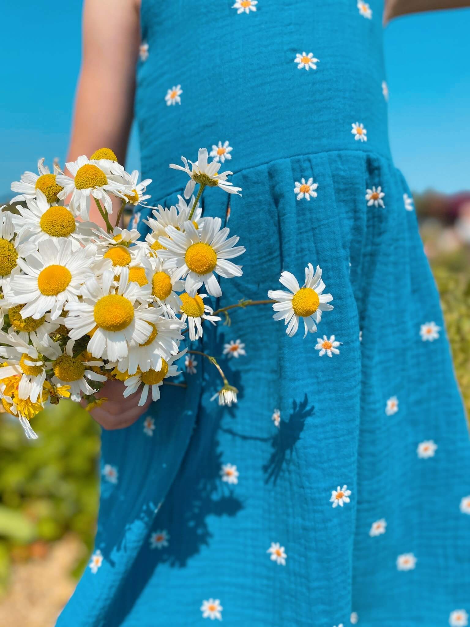 Mädchen in einem Kleid in blau mit weißen Gänseblümchen aus Musselinstoff von LA.LUKA Berlin, hält einen Strauß Gänseblumen in der Hand.