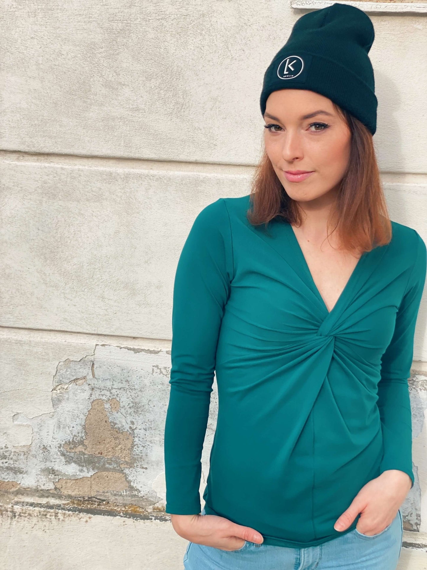 Frau in einem dunkelgrünen Knotenshirt aus Baumwolljersey mit langen Ärmeln und schwarze Mütze mit Logo von LA.LUKA Berlin.