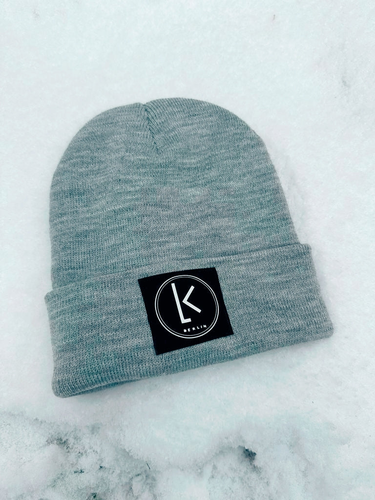 Graue Mütze mit dem Logo von LA.LUKA Berin, liegt zm Schnee.