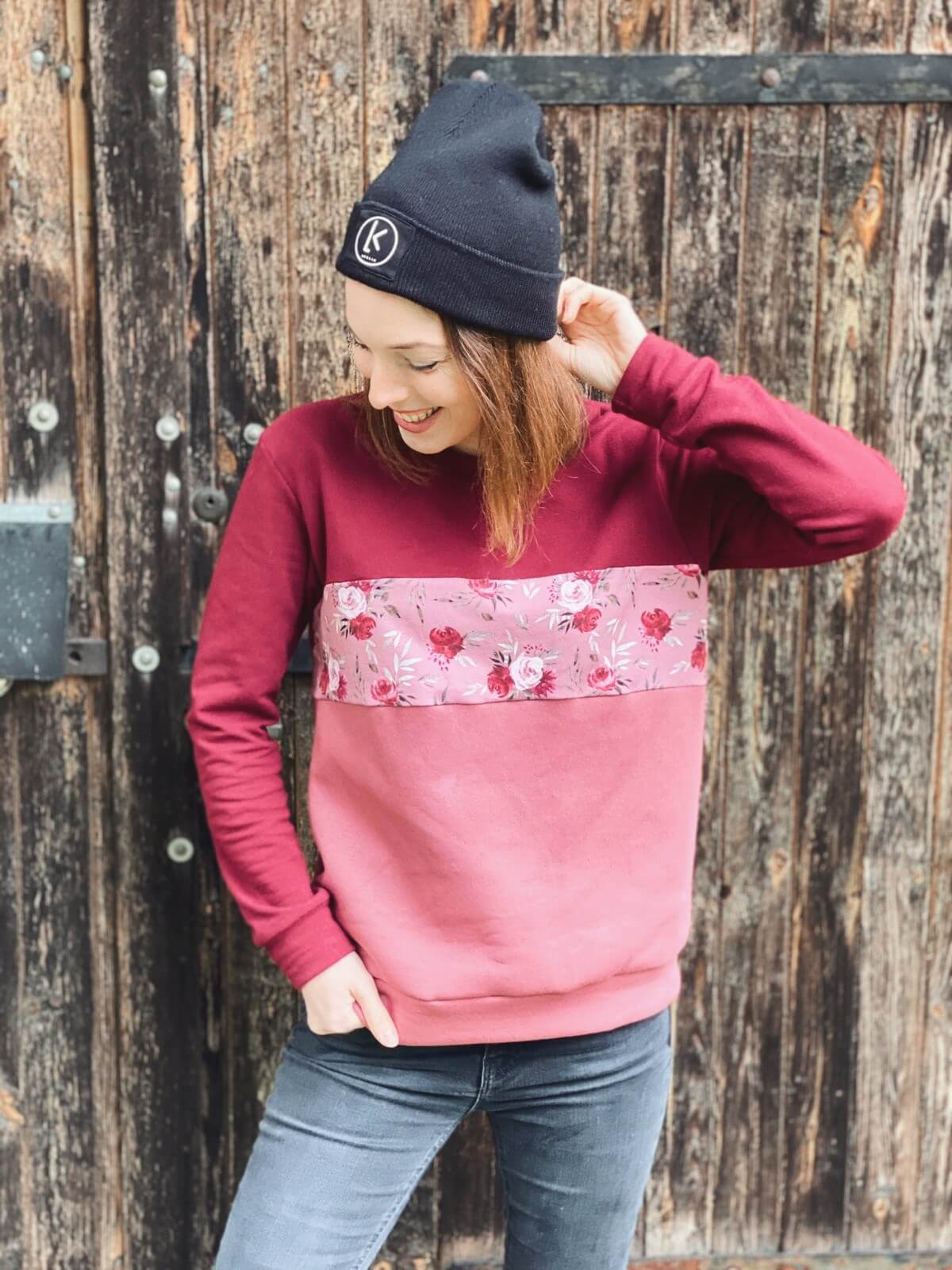 Lachende Frau mit schwarzer Mütze und rosé farbenen Pullover mit Blumenprint von LA.LUKA Berlin vor einer alten Holztür.