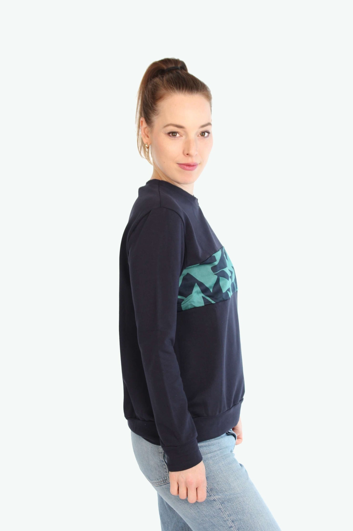 Eine seitlich stehende Frau, in einem dunkelblauen Pullover mit grün-blauen Muster auf der Brustpartie.