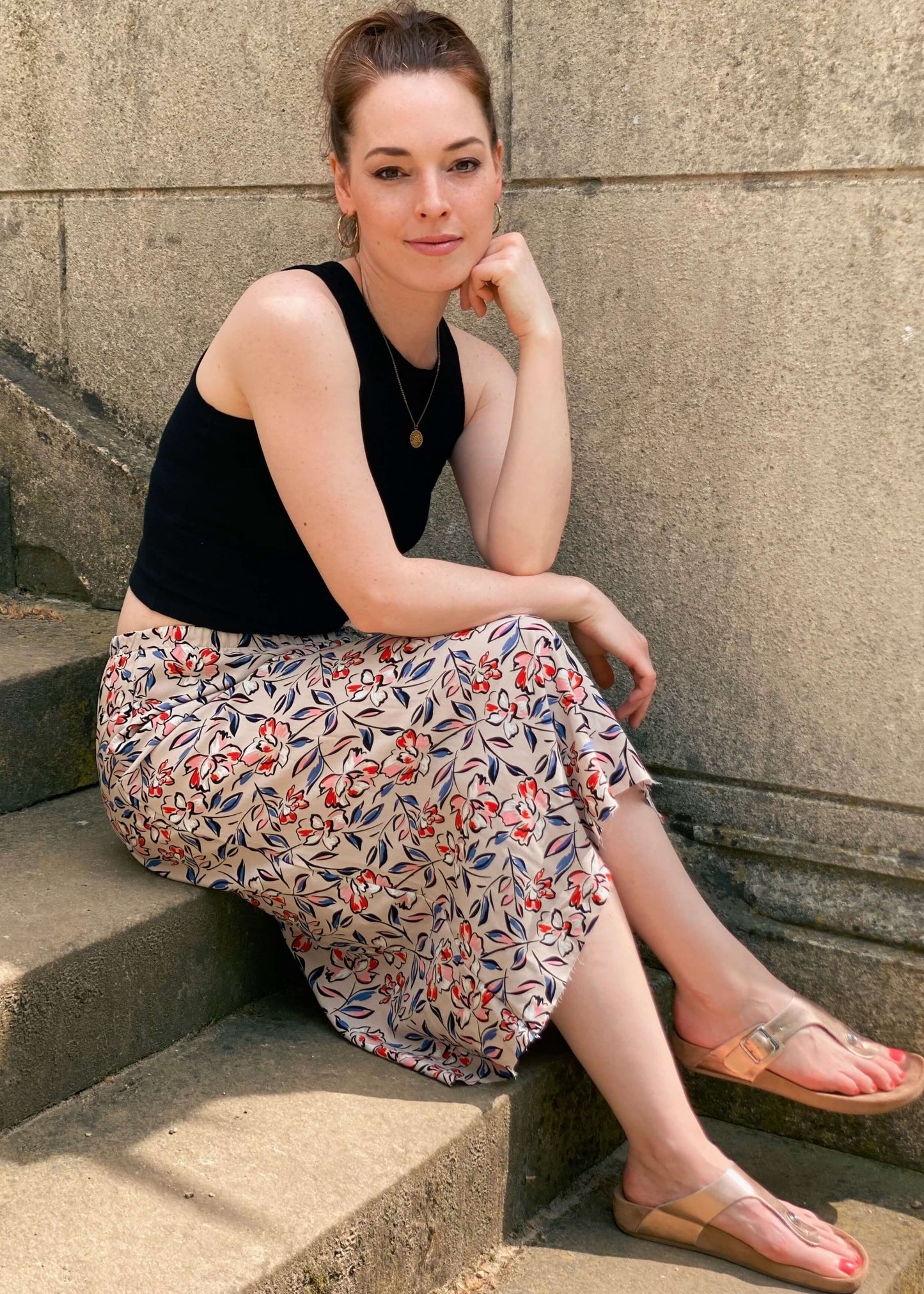 Frau, sitzt auf einer Treppe in einem schwarzen Top und einem Midirock in beige mit bunten Blumen von LA.LUKA Berlin.
