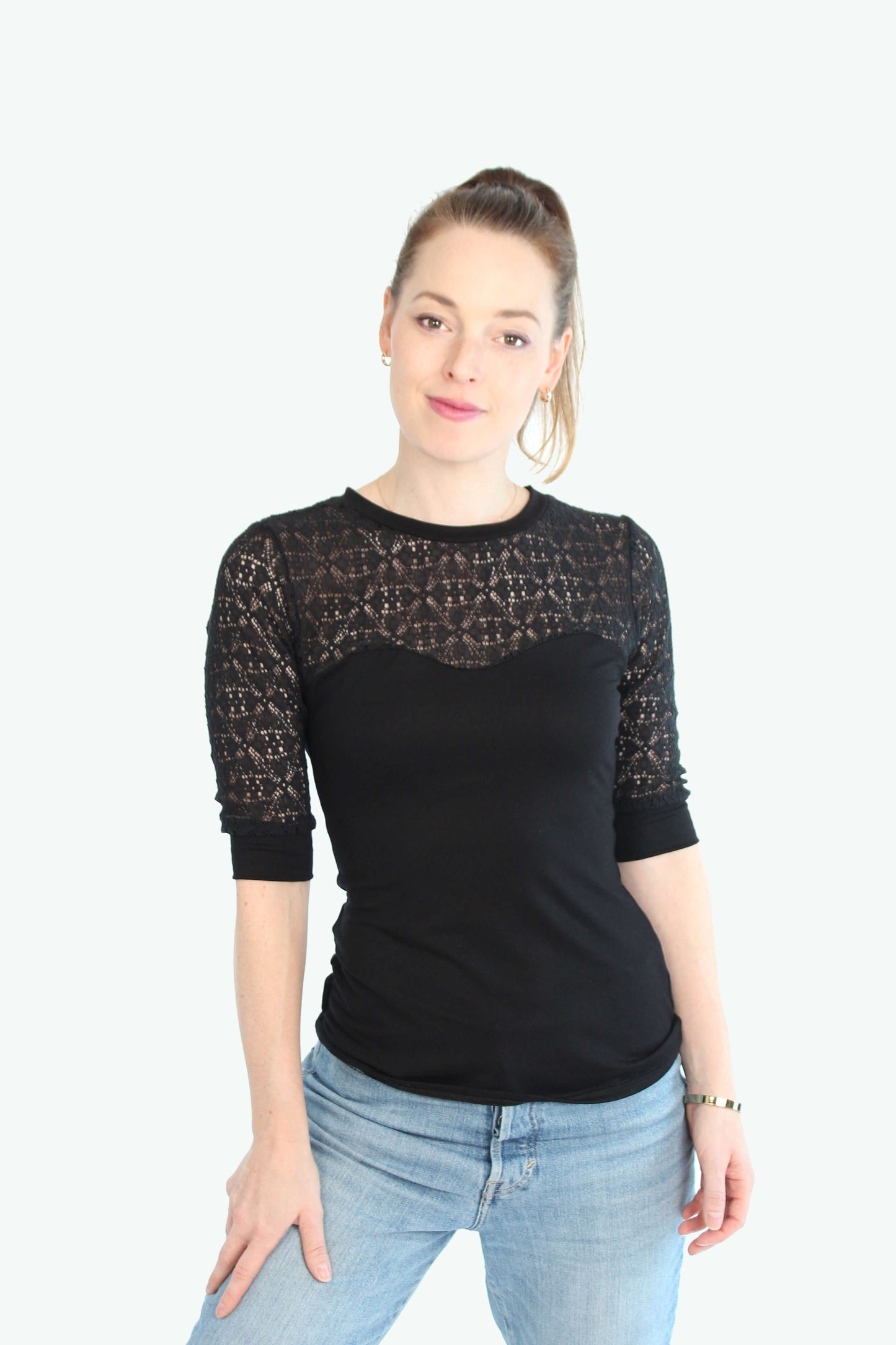 Eine Frau trägt ein schwarzes Spitzentop mit halblangen Ärmeln. Das Oberteil hat einen figurbetonten Schnitt, eine transparente Spitzenpartie an Schultern und Dekolleté sowie eine elegante Optik. Kombiniert wird das Top mit einer hellblauen Jeans.