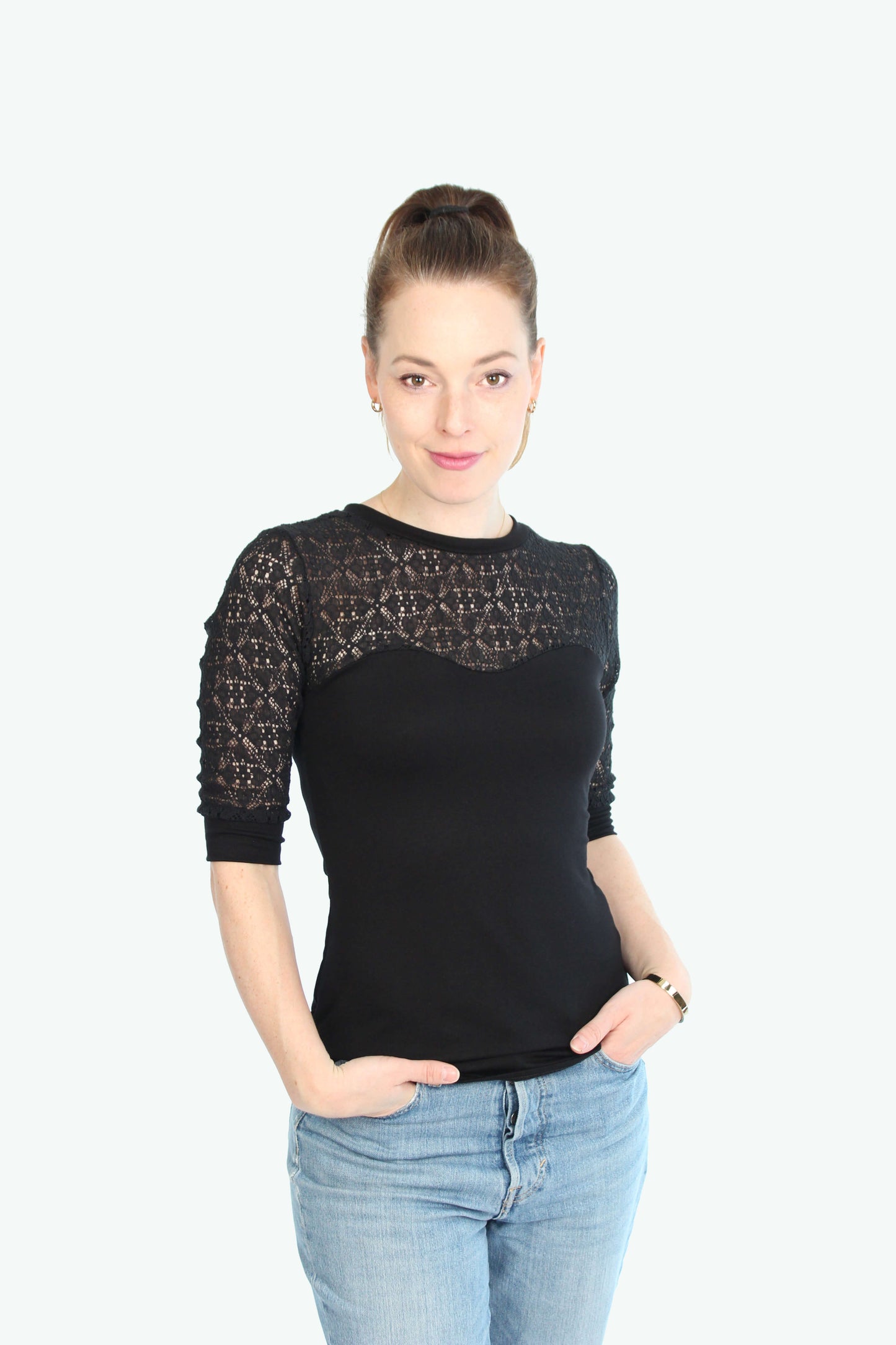 Eine Frau trägt ein schwarzes Spitzentop mit halblangen Ärmeln und transparenter Spitze an Schultern und Dekolleté, kombiniert mit einer hellblauen Jeans.