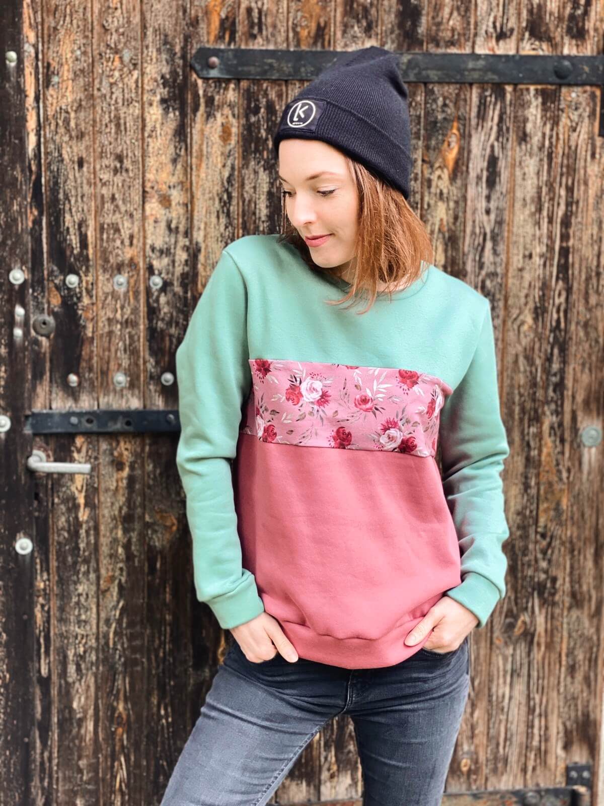 Frau, trägt einen Pullover aus Baumwolle in mint und rosé mit Blumenprint dazu eine schwarze Mütze mit dem Logo von LA.LUKA Berlin.
