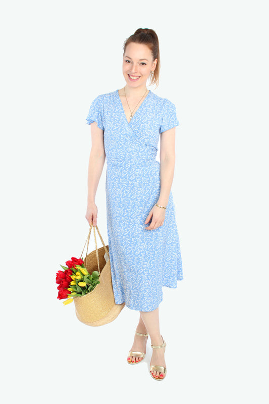 Frau in einem Sommerkleid in Midilänge zum wickeln in hellblau mit weißen Blümchenmuster aus Viskosestoff von LA.LUKA Berlin, trägt eine Korbtasche mit Tulpen. 