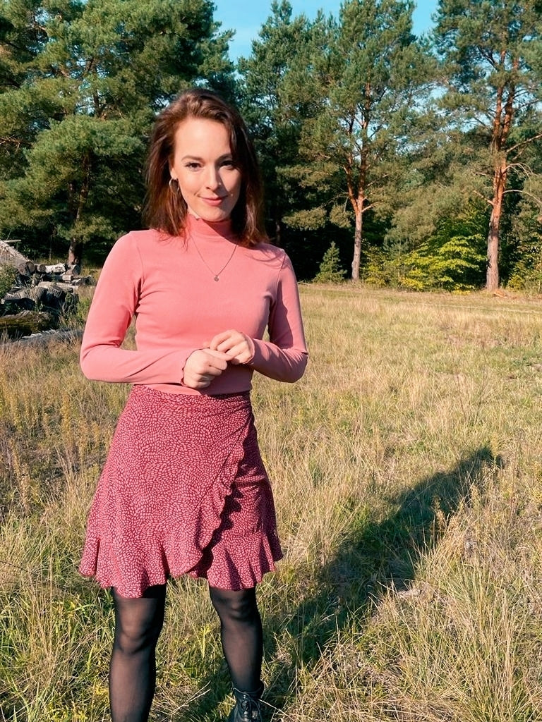 Frau in einem Rollkragenshirt und kurzen Wickelrock in bordeaux mit Pünktchen in rosé aus Viskosestoff von LA.LUKA Berlin, steht auf einer Lichtung im Wald.