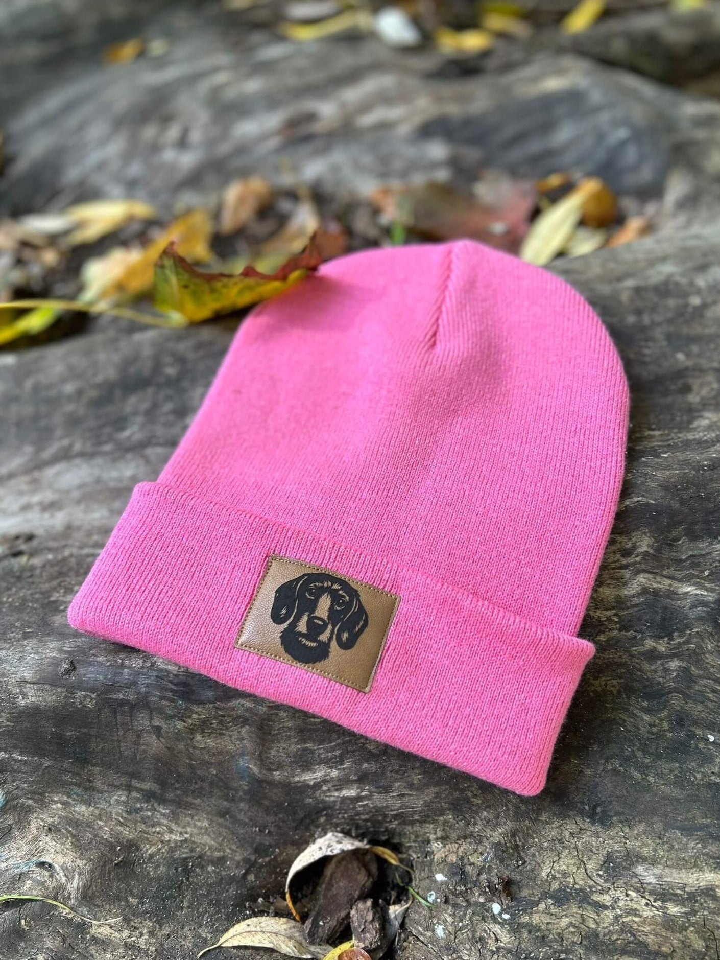 „Pinkfarbene Beanie-Mütze von LA.LUKA Berlin mit Kunstlederetikett und geprägtem Rauhaardackel-Motiv, platziert auf rustikalem Holzuntergrund mit bunten Herbstblättern.