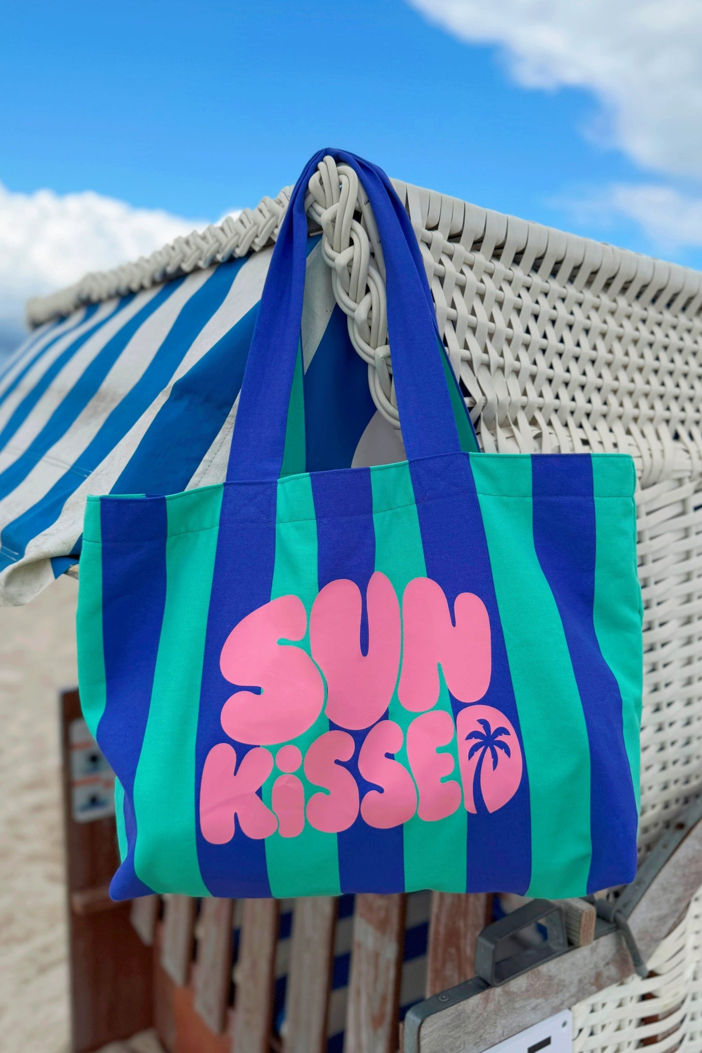 Große gestreifte Strandtasche aus Canvas in Türkis und Blau mit dekorativem rosa Foliendruck an einem Strandkorb am Strand.