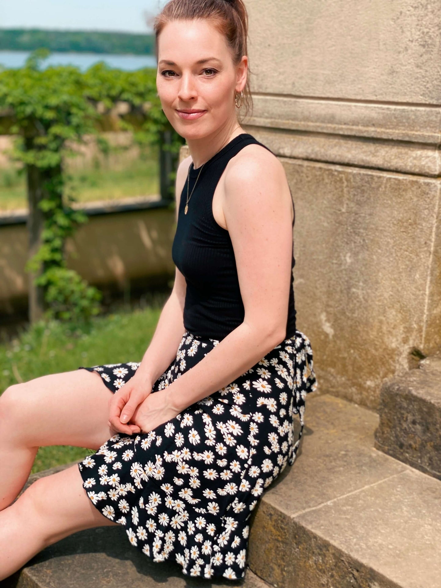 Frau in einem schwarzen Wickelrock mit weißen Gänseblümchen aus Viskose von LA.LUKA Berlin, sitzt auf einer Treppe.