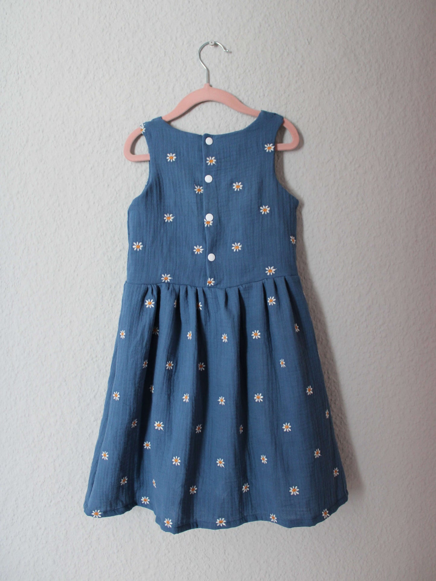 Kinderkleid in jeansblau mit weißen Gänseblümchen aus Musselinstoff von LA.LUKA Berlin auf einem Kleiderbügel in der Rückansicht.