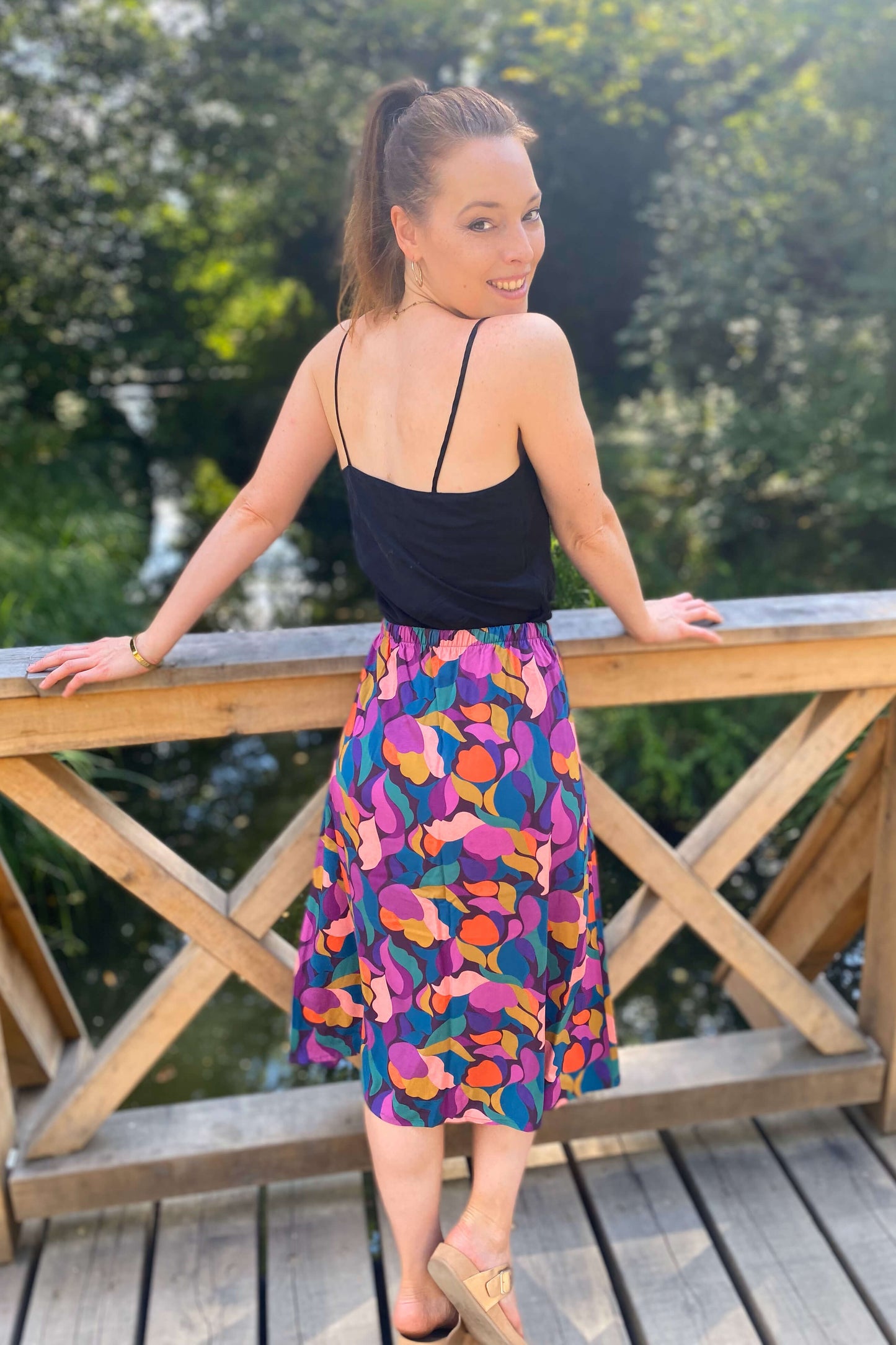 Frau auf Holzbrücke von hinten mit farbenfrohem Midirock in Lila, Orange und Pink von LA.LUKA Berlin – sommerlicher Look in der Natur.