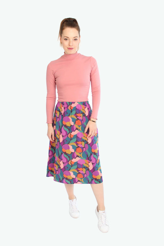 Frau trägt bunten Midirock mit abstraktem Muster in Pink, Lila und Orange von LA.LUKA Berlin – kombiniert mit rosa Rollkragen-Top und weißen Sneakern.