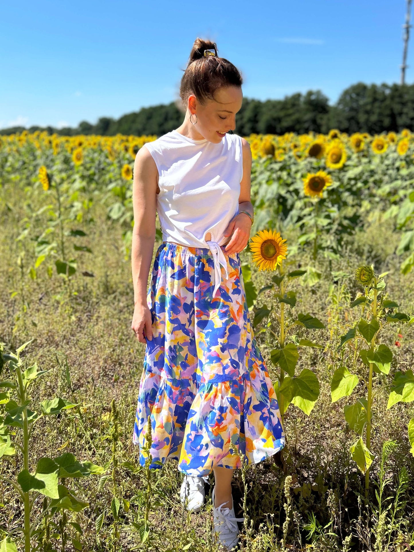 Frau steht im Sonnenblumenfeld und trägt einen bunten Sommerrock in Midilänge mit floralem Muster von LA.LUKA Berlin, kombiniert mit einem weißen, ärmellosen Knotentop – perfekter Look für den Sommer 2025.