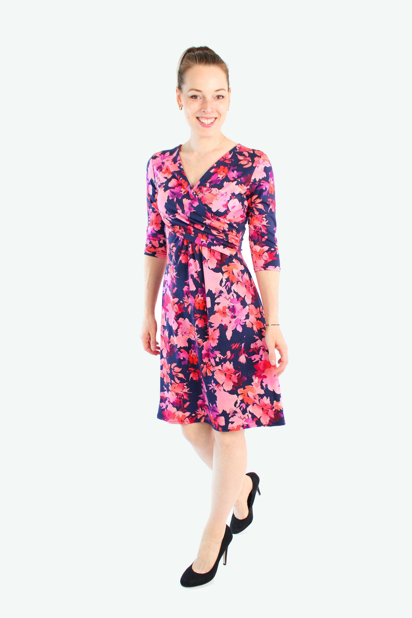 Model trägt dunkelblaues Kleid mit pinken Blumenmuster. Durch die Wickeloptik wird die feminine Silhouette betont, kombiniert mit schwarzen Pumps.
