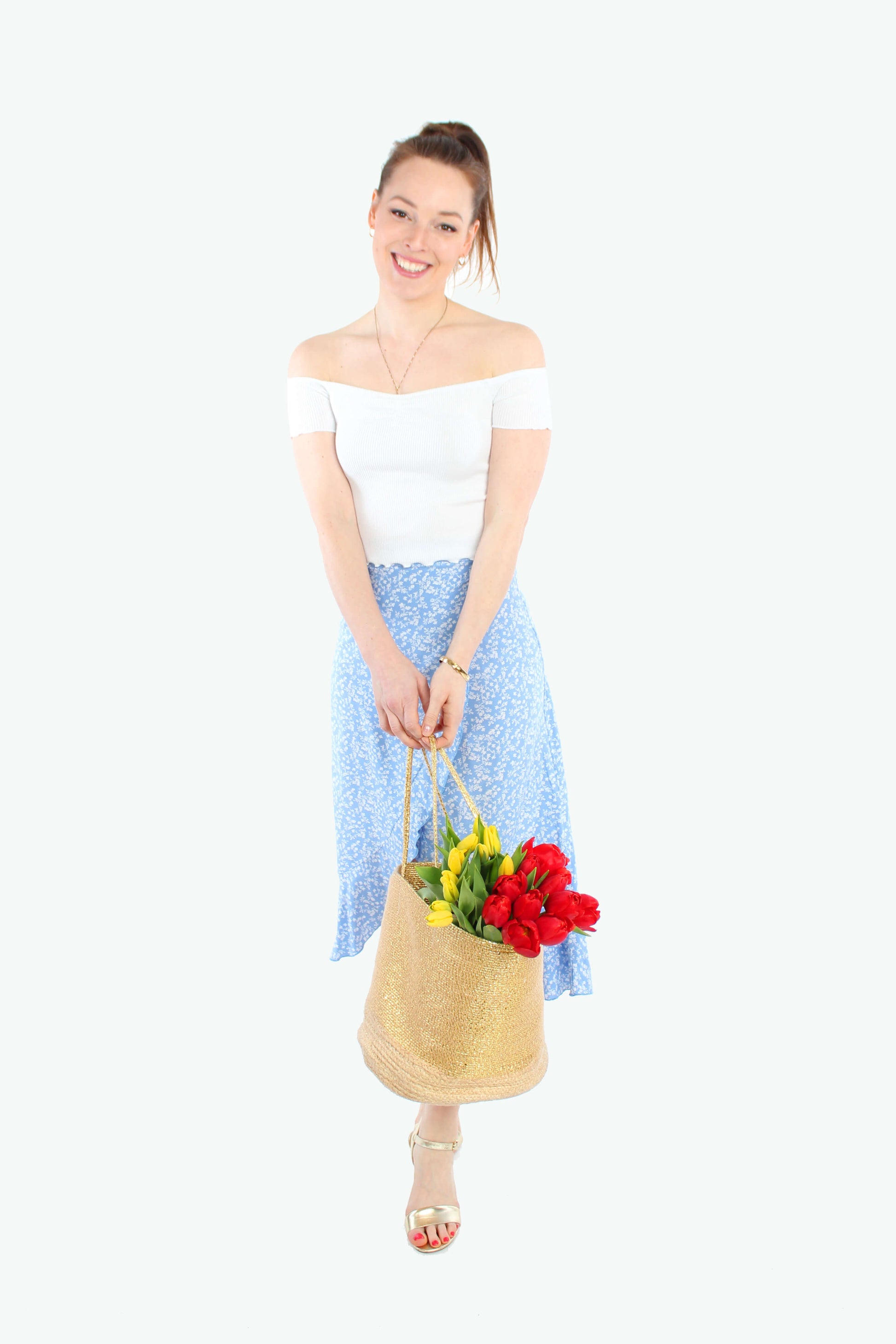 Frau, in einem schwingenden, leichten Wickelrock in Midilänge in hellblau mit weißen Blümchen und Volant von LA.LUKA Berlin, sie hält eine Tasche mit Blumen in der Hand.
