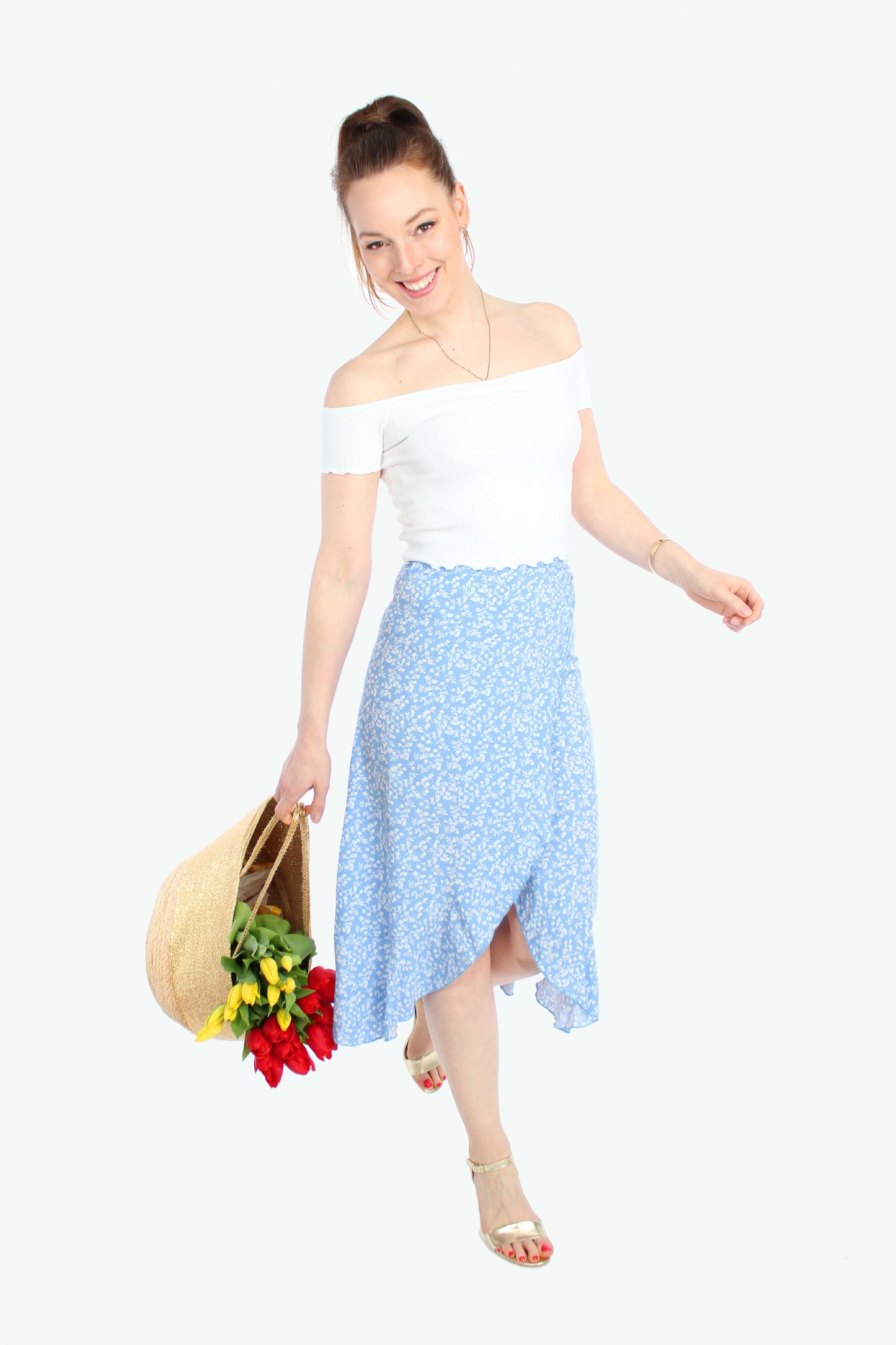 Frau, in einem sommerlichen, leichten Wickelrock in Midilänge in hellblau mit weißen Blümchen und Volant von LA.LUKA Berlin, sie hält eine Tasche mit Blumen in der Hand.