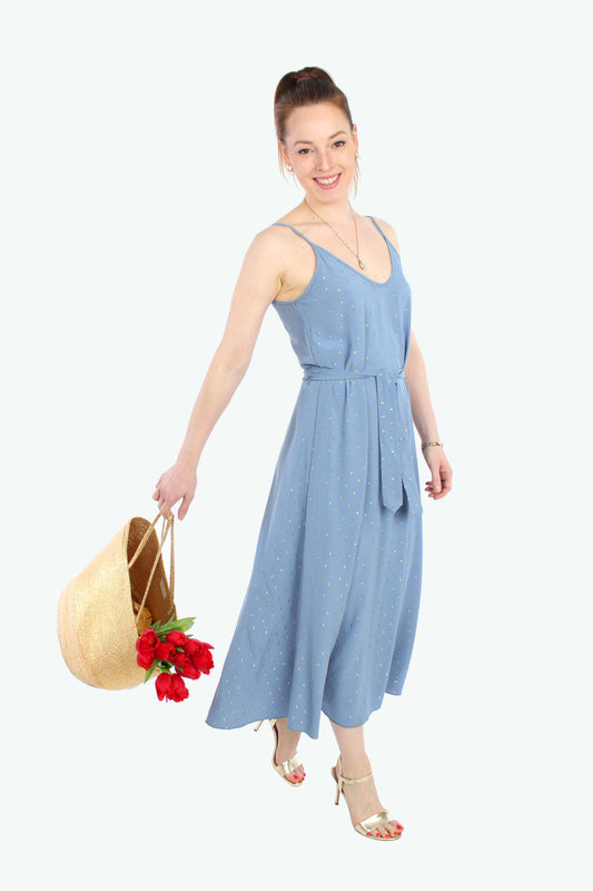 Frau, in einem hellblauen Sommerkleid in Midilänge mit goldenen Pünktchen mit Spaghettiträgern aus leichtem Viskosestoff von LA.LUKA Berlin, in der Hand eine Korbtasche mit Tulpen. 
