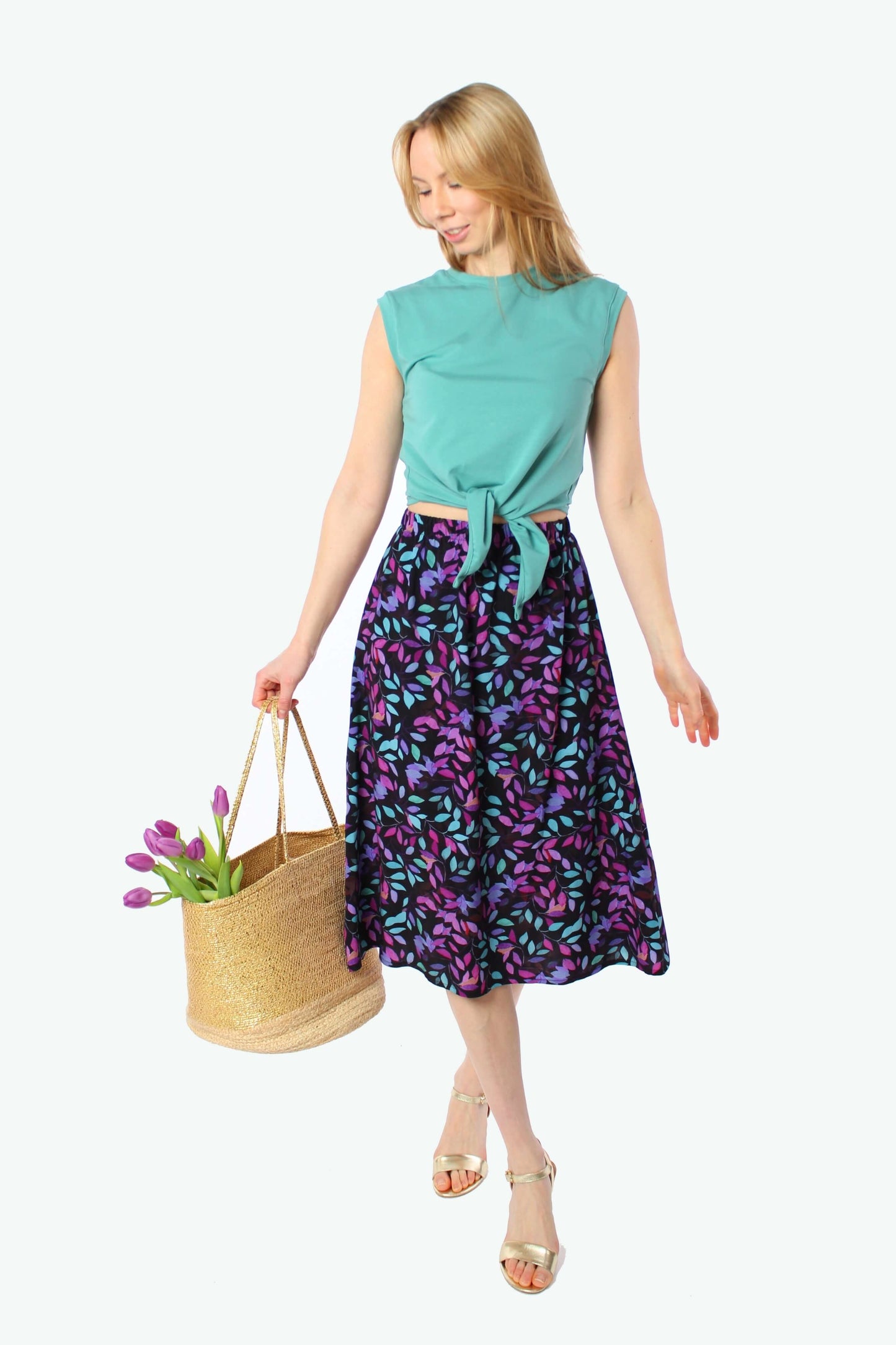 Model mit langen blonden Haaren trägt fließenden Midirock in Schwarz mit lila und türkisem Muster, elastischer Gummibund, sommerlich kombiniert mit Top und Sandalen.