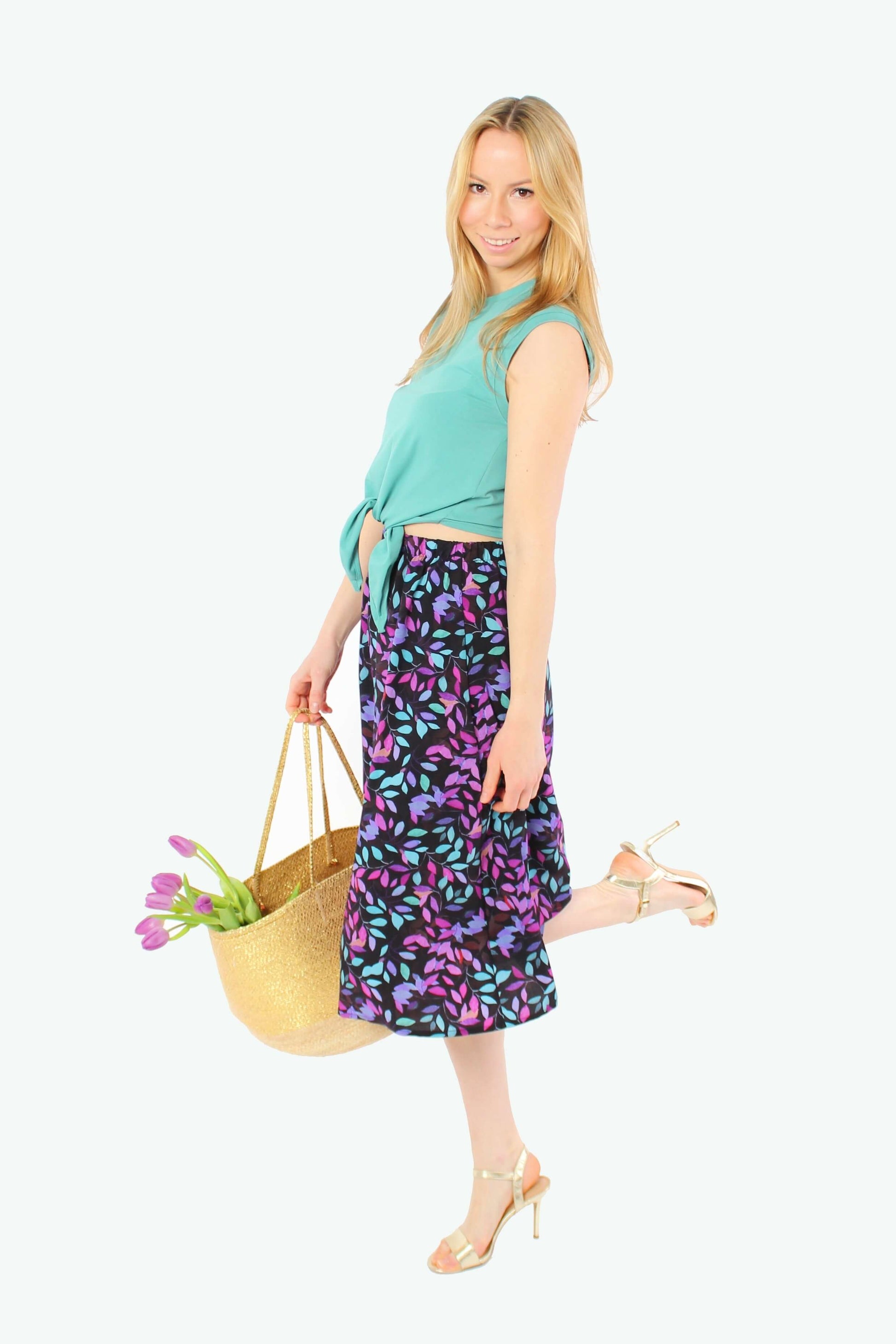 Blondes Model in Seitenansicht mit schwarzem Sommerrock in Midi-Länge aus Viskose, lila-türkises Allover-Muster, elastischer Bund, kombiniert mit grünem Top und goldenen High Heels.