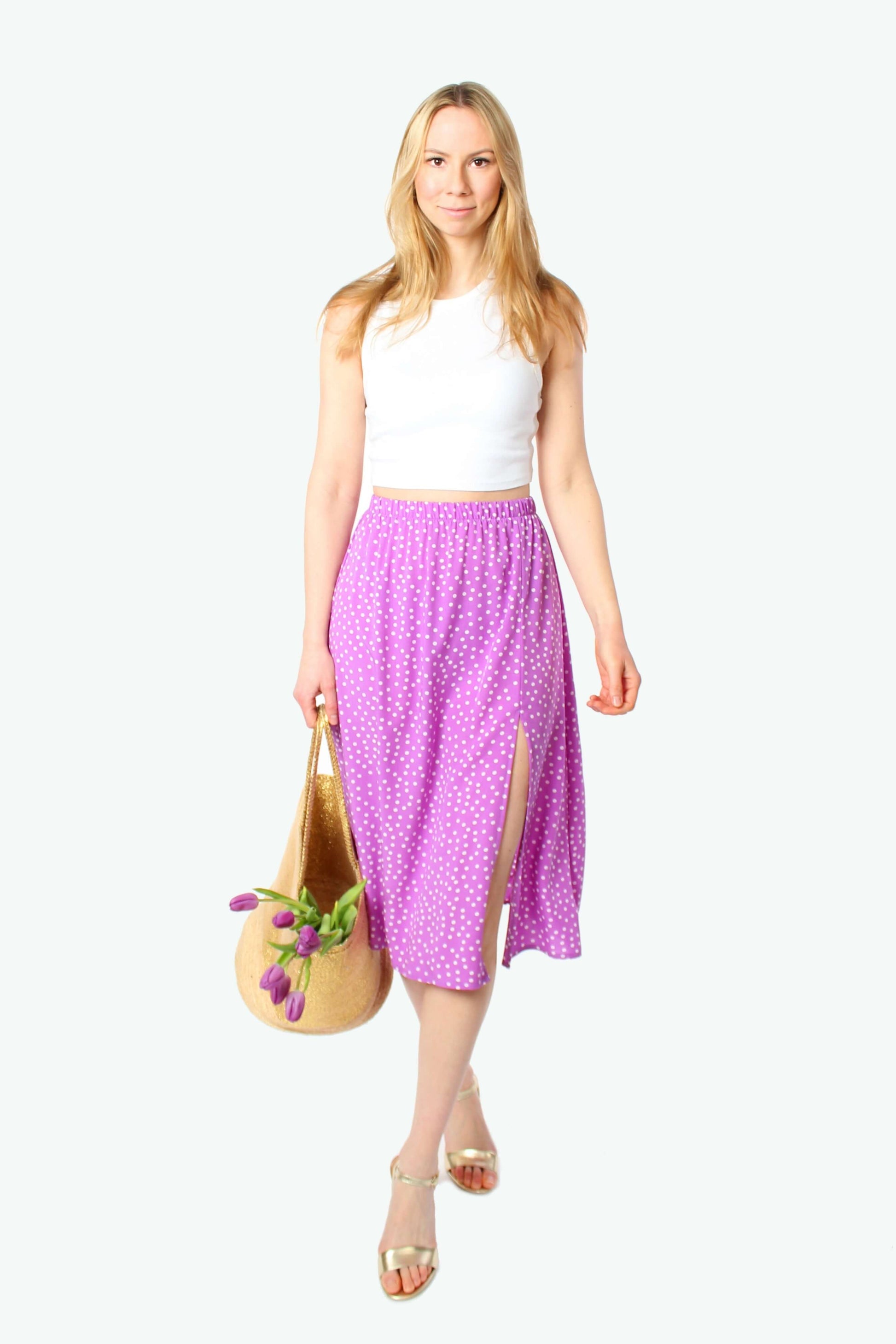 Blondes Model trägt lila Midirock mit weißen Punkten aus 100% Viskose, elastischer Bund und seitlicher Schlitz, kombiniert mit weißem Top und Sandalen.