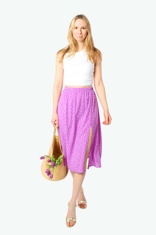 Blondes Model trägt lila Midirock mit weißen Punkten aus 100% Viskose, elastischer Bund und seitlicher Schlitz, kombiniert mit weißem Top und Sandalen.
