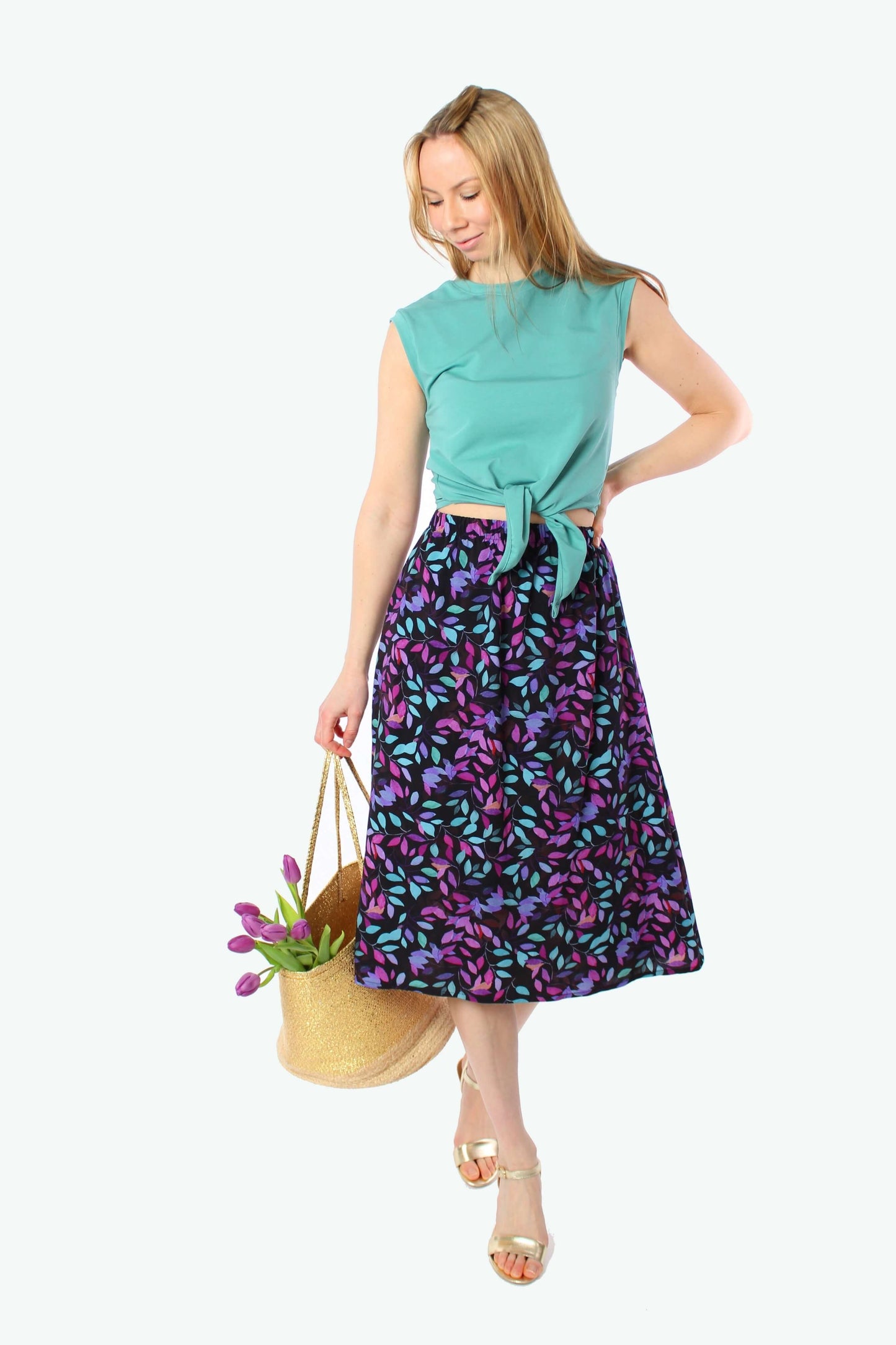 Blondes Model trägt einen schwarzen Midirock aus 100% Viskose mit lila und türkisfarbenem Blattmuster, kombiniert mit grünem, geknotetem Top, goldenen Sandalen und einer geflochtenen Strohtasche mit lila Tulpen.