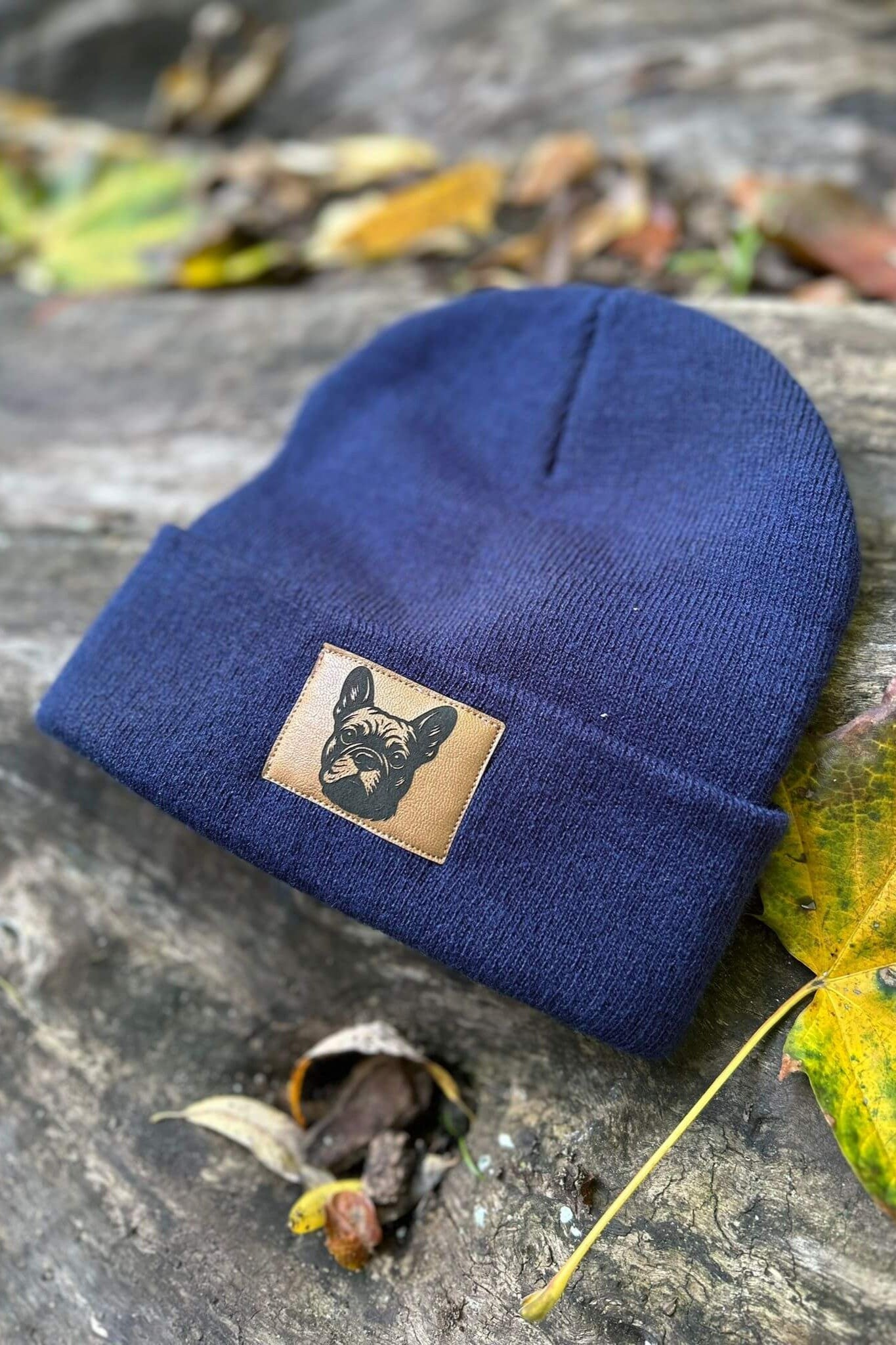 Dunkelblaue Mütze von LA.LUKA Berlin mit Kunstlederpatch und Motiv einer Französischen Bulldogge, liegend auf Holz mit Herbstblättern – stilvolles Hunde-Accessoire für kalte Tage.
