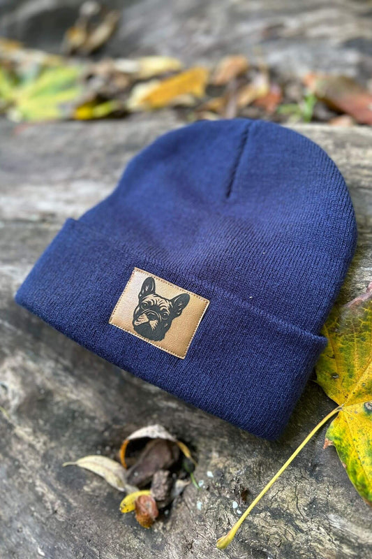 Dunkelblaue Mütze von LA.LUKA Berlin mit Kunstlederpatch und Motiv einer Französischen Bulldogge, liegend auf Holz mit Herbstblättern – stilvolles Hunde-Accessoire für kalte Tage.