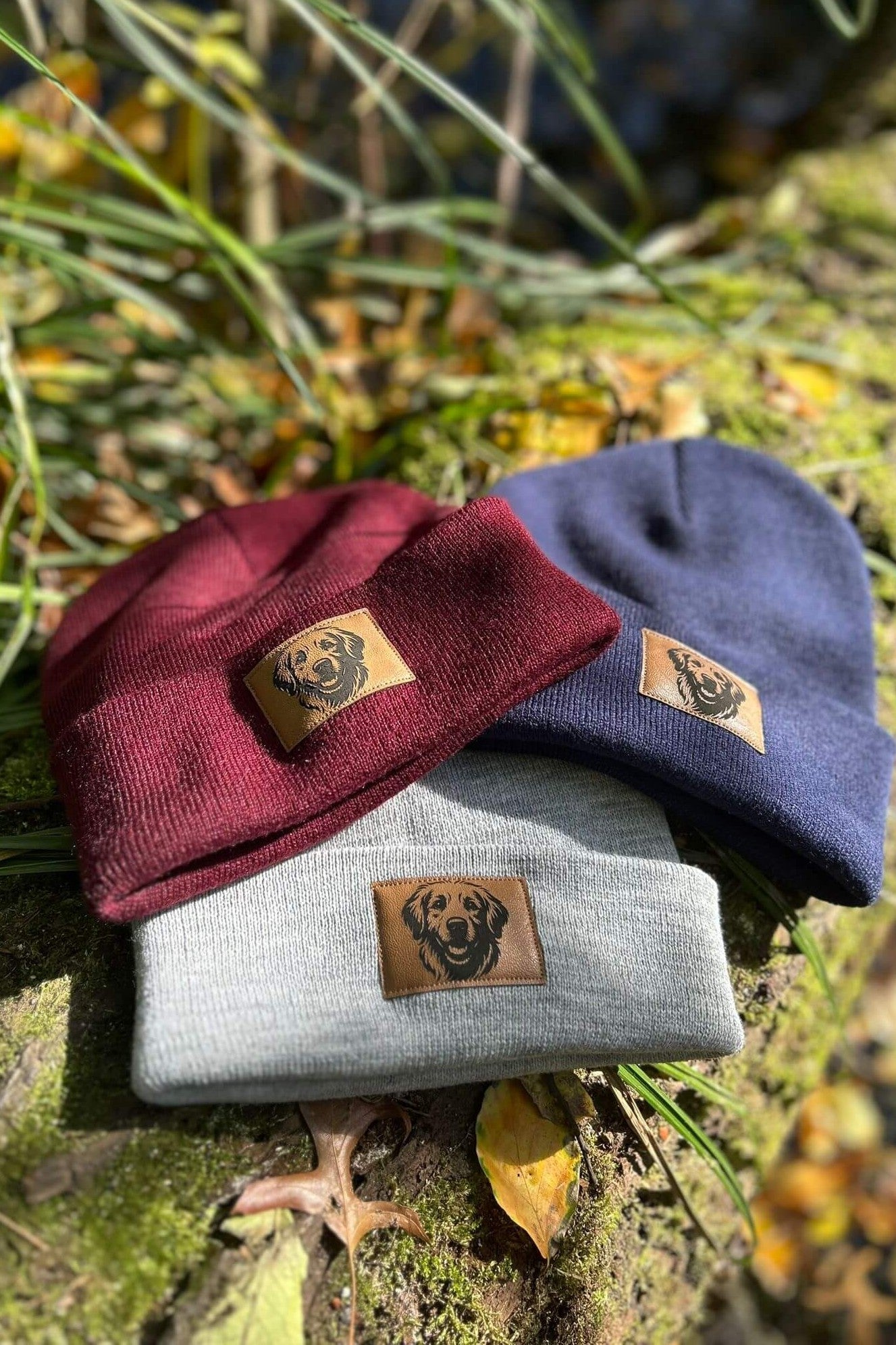 Drei Mützen in Bordeaux, Grau und Dunkelblau von LA.LUKA Berlin mit Kunstleder-Patch und Labrador-Motiv, arrangiert auf Moos und Herbstlaub – warme Beanies für Hundefans.
