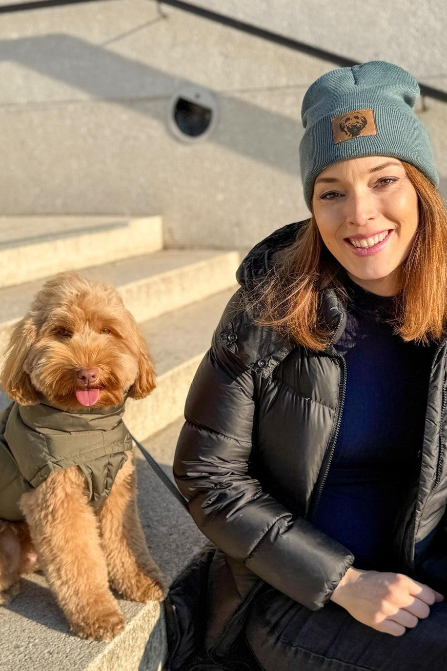 Frau sitzt mit einem kleinen flauschigen Hund auf Treppenstufen und trägt eine salbeigrüne Beanie mit Labradoodle-Patch – warme Wintermütze für Hundefreunde.