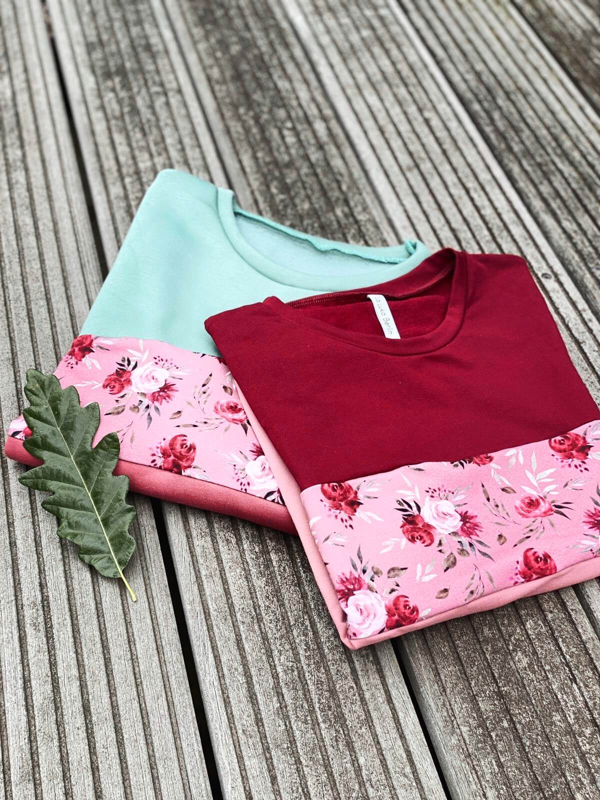 Zwei Pullover in rose mit Blumenmuster, liegen zusammengelegt auf Holzboden.