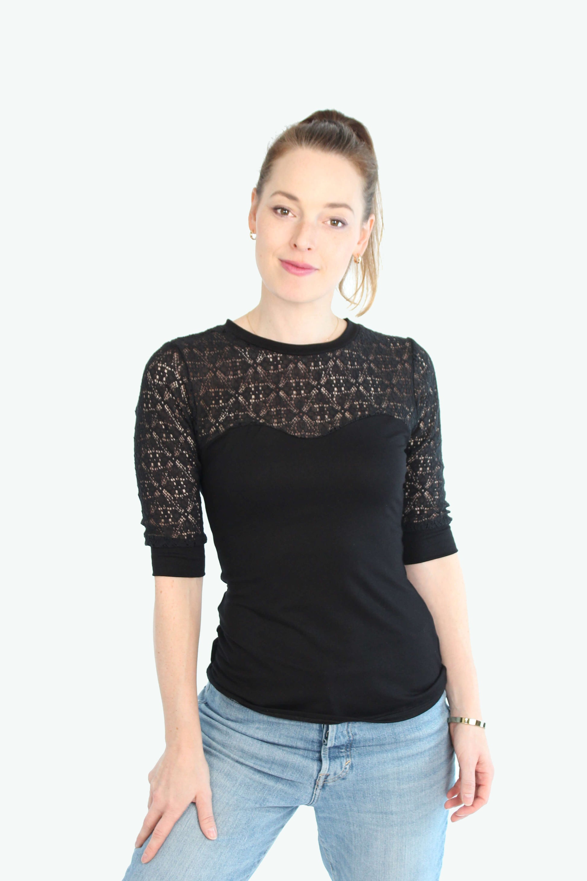Eine Frau trägt ein schwarzes Spitzentop mit halblangen Ärmeln. Das Oberteil hat einen figurbetonten Schnitt, eine transparente Spitzenpartie an Schultern und Dekolleté sowie eine elegante Optik. Kombiniert wird das Top mit einer hellblauen Jeans.