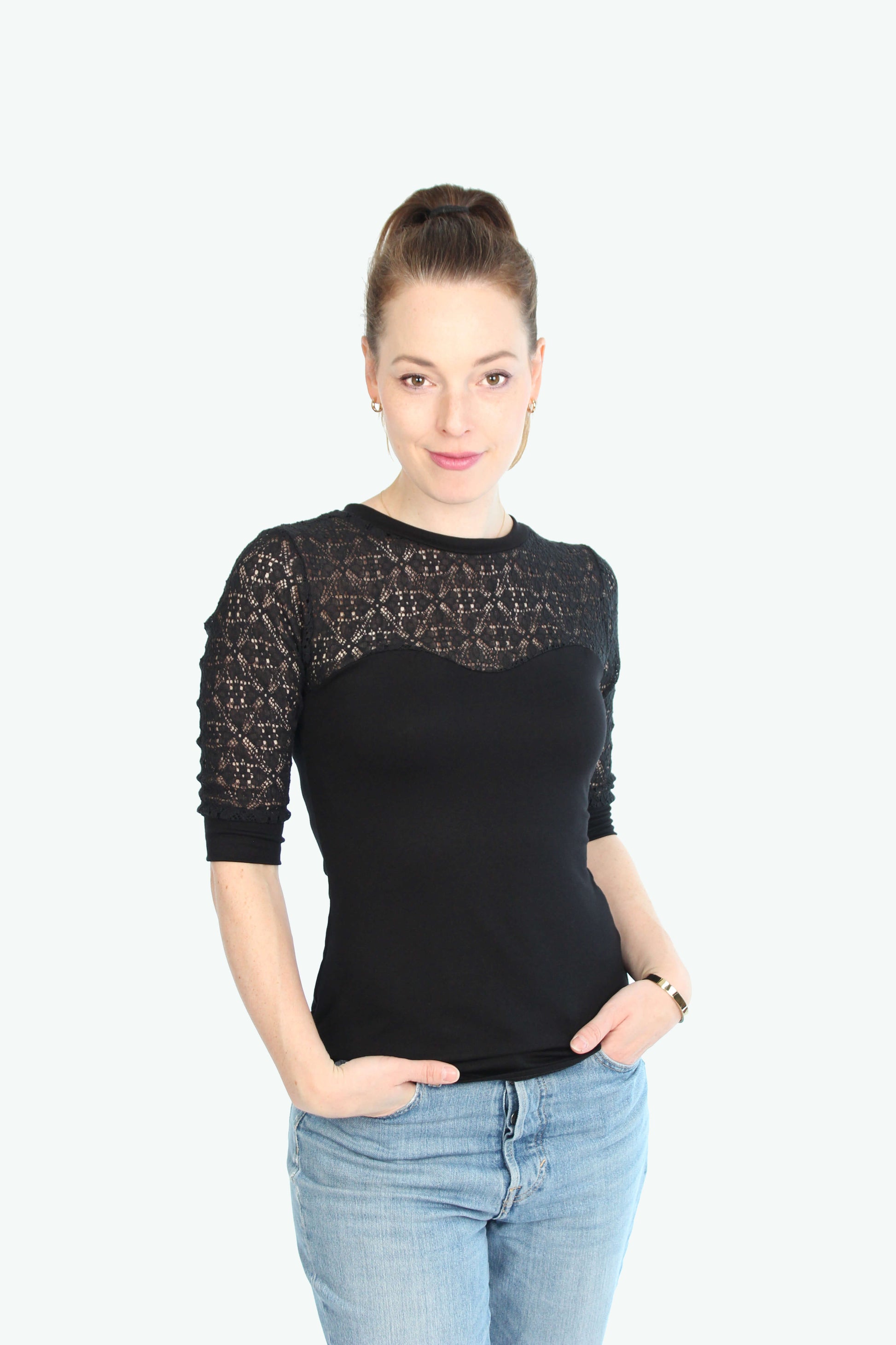 Eine Frau trägt ein schwarzes Spitzentop mit halblangen Ärmeln und transparenter Spitze an Schultern und Dekolleté, kombiniert mit einer hellblauen Jeans.