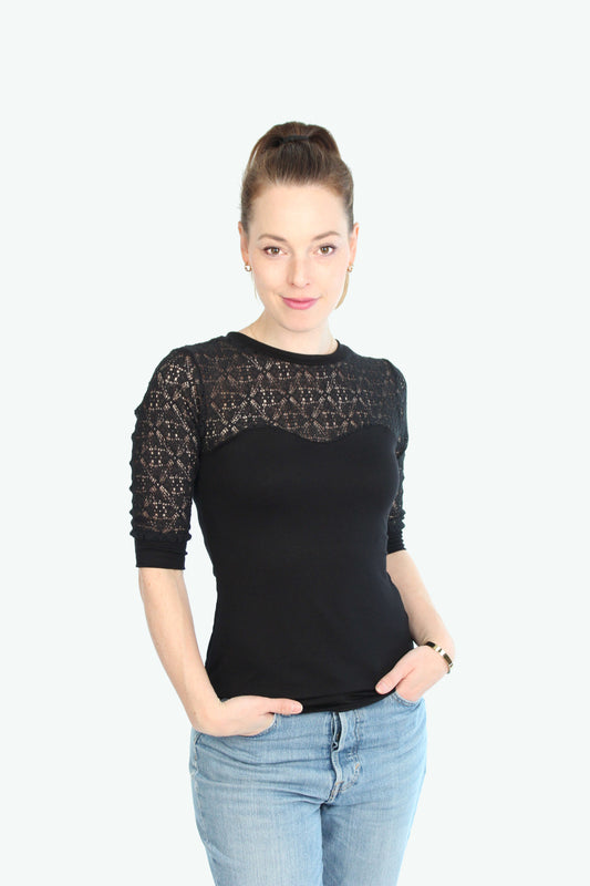 Eine Frau trägt ein schwarzes Spitzentop mit halblangen Ärmeln und transparenter Spitze an Schultern und Dekolleté, kombiniert mit einer hellblauen Jeans.