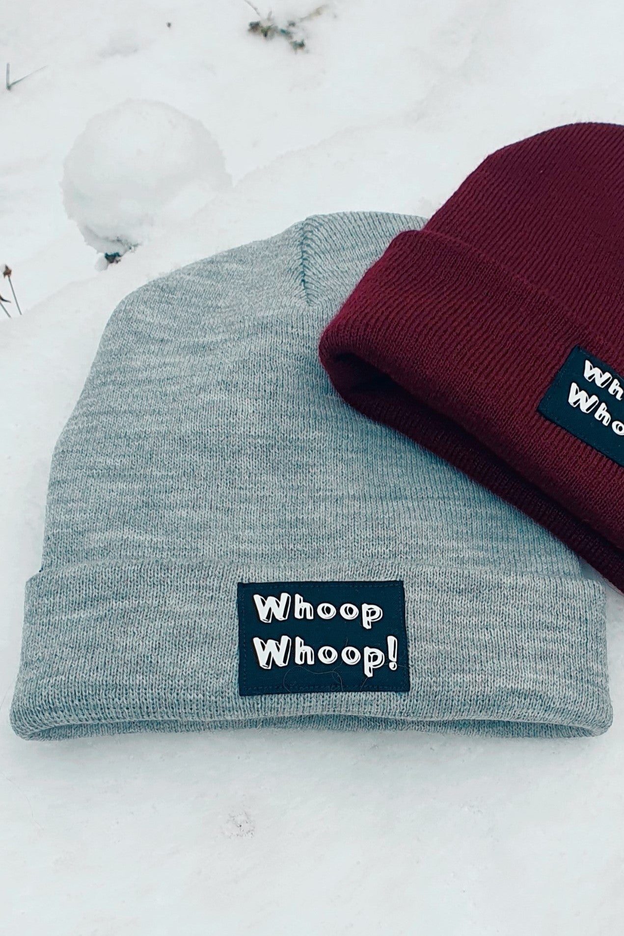 Zwei Mützen mit Whoop Whoop! Motiv in grau und bordeaux von LA.LUKA Berlin, liegen im Schnee.