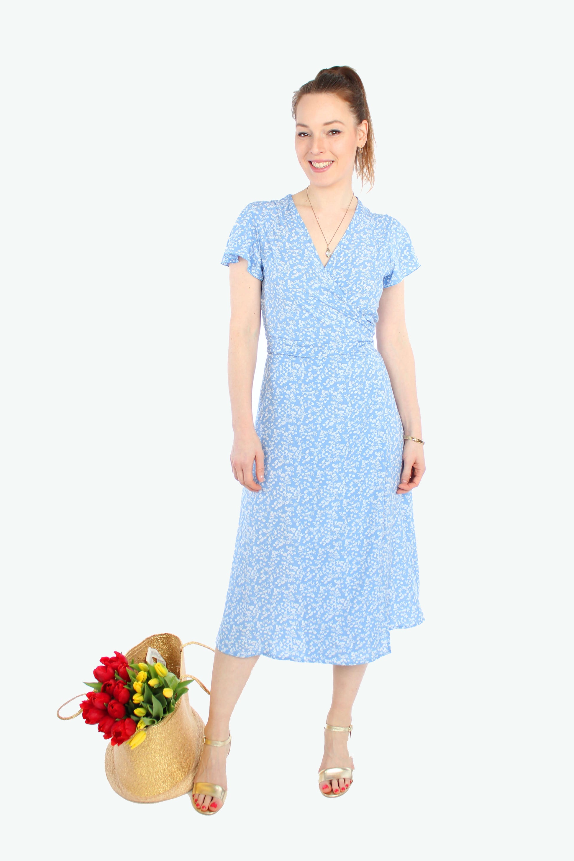 Frau in einem Sommerkleid in Midilänge zum wickeln in hellblau mit weißen Blümchenmuster aus leichtem Viskosestoff von LA.LUKA Berlin, neben ihr steht eine Korbtasche mit Tulpen. 