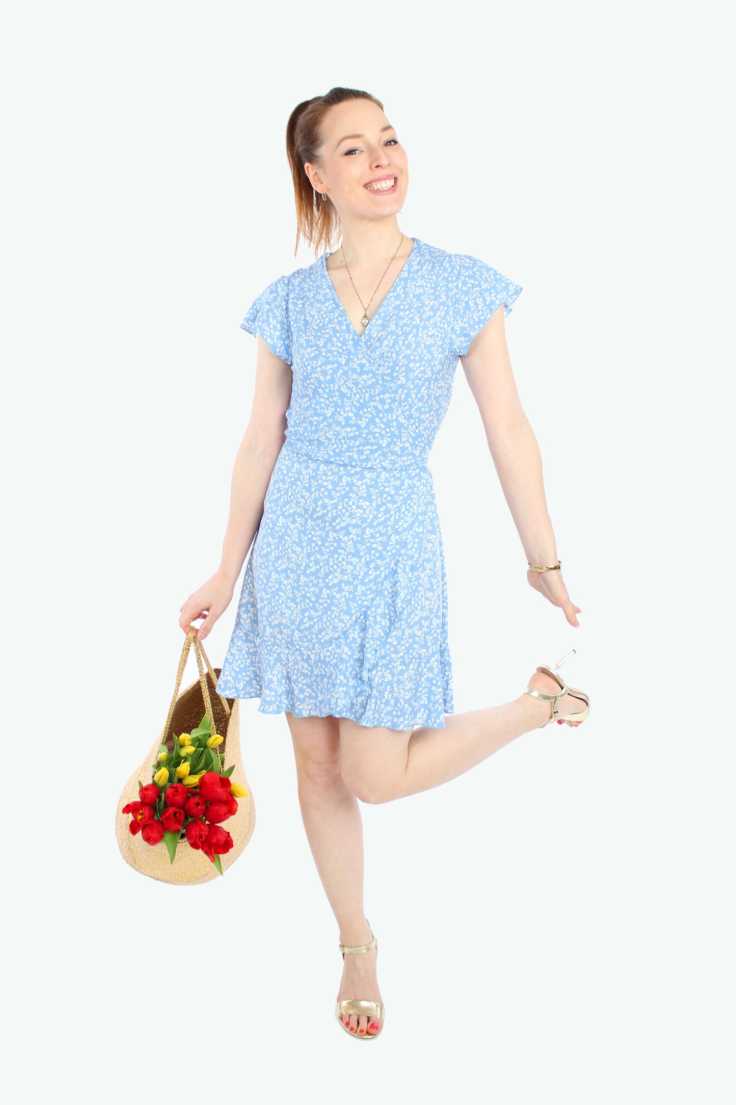Lachende Frau in einem Sommerkleid in Minilänge zum wickeln in hellblau mit weißen Blümchenmuster aus leichtem Viskosestoff von LA.LUKA Berlin, trägt eine Korbtasche mit Tulpen. 