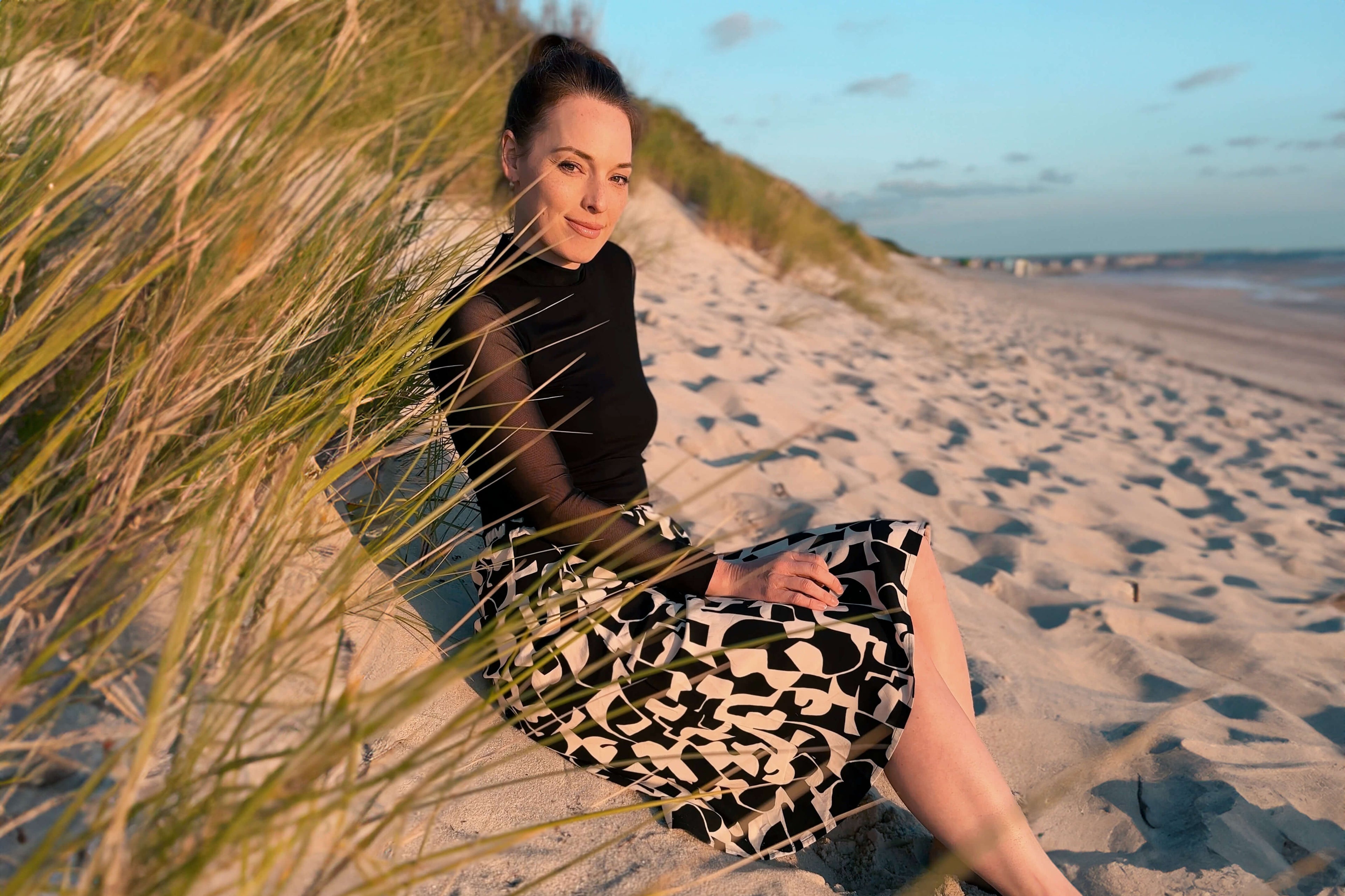 Frau, sitzt am Strand, nah der Dünen. Sie blickt in die Kamera und trägt einen schwar, weiß gemusterten Midirock uns ein schwarzen Oberteil.
