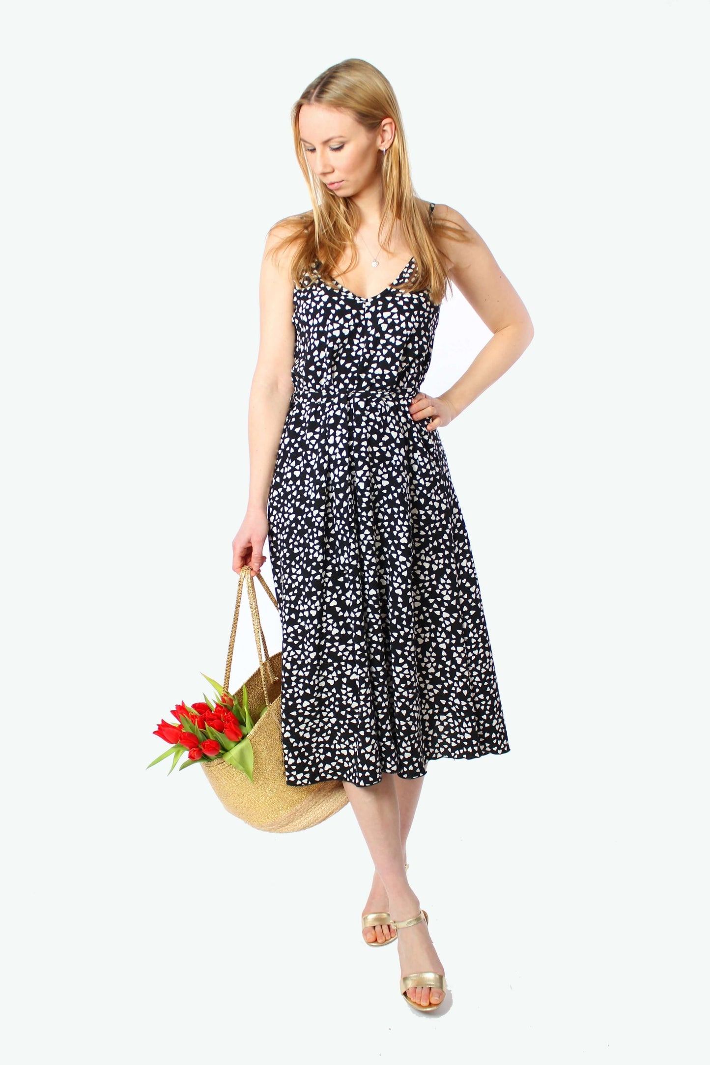 Frau trägt ein schwarzes Midikleid mit kleinen weißen Herzen, schmale Träger und V-Ausschnitt, das Kleid reicht bis unter das Knie und fällt locker. Kombiniert mit goldenen Sandalen und großer Basttasche mit roten Tulpen.