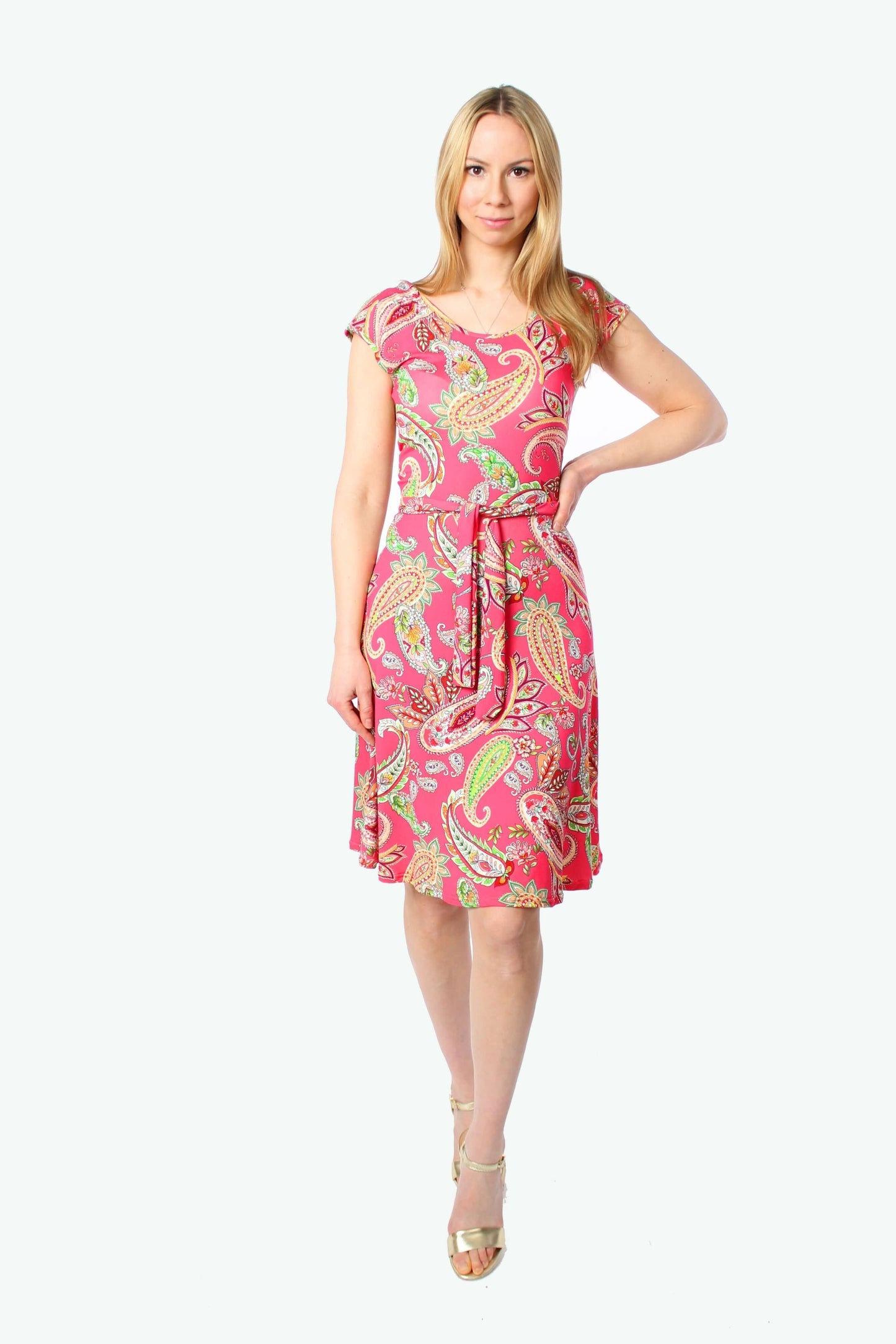 Blonde Frau steht frontal und trägt ein pinkes Sommerkleid mit Paisley-Muster, knielanges Jerseykleid mit kurzen Ärmeln und Bindegürtel.