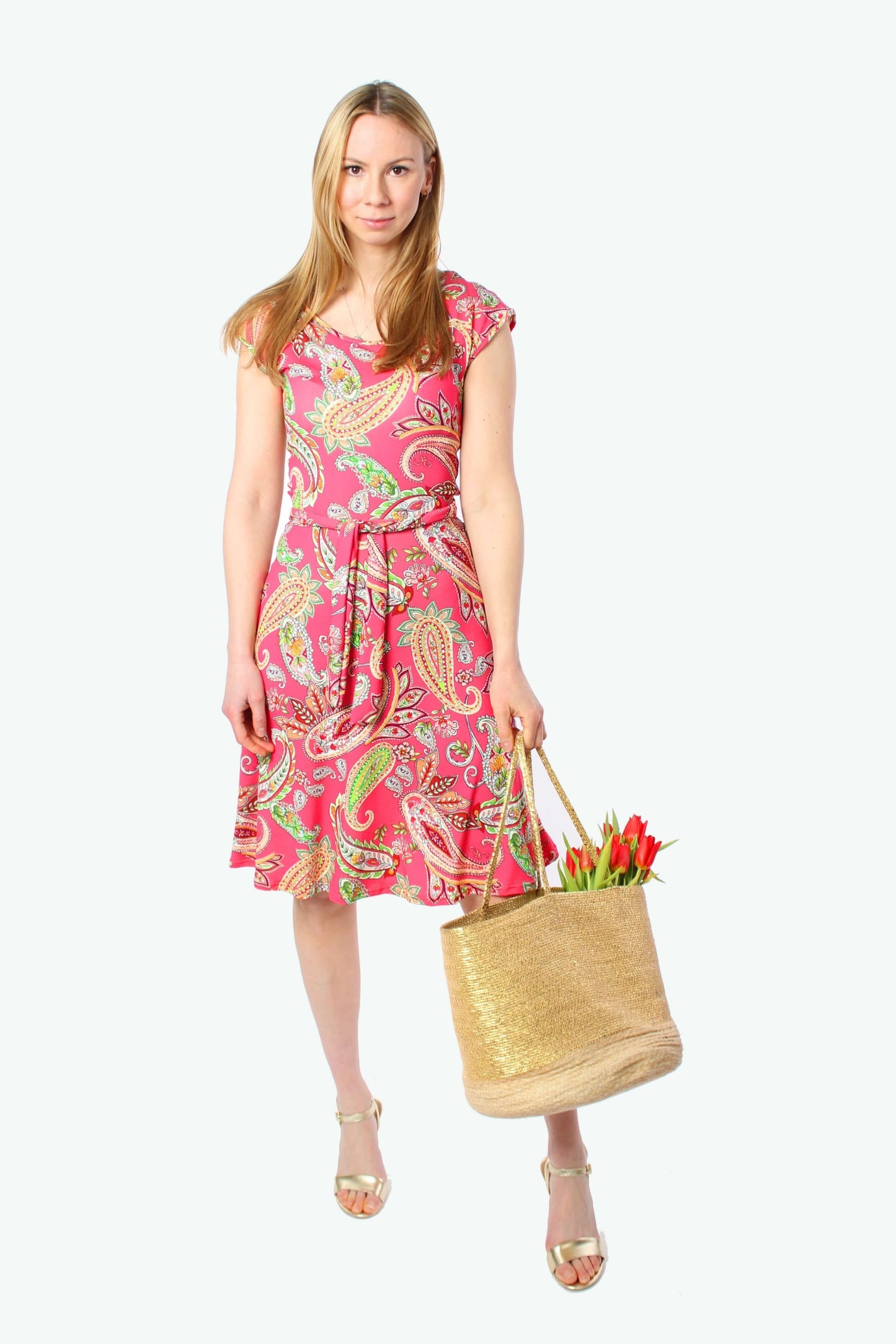 Blonde Frau im pinken Paisley Sommerkleid von LA.LUKA Berlin aus Jersey, knielang mit Bindegürtel, sie hält eine goldene Tasche mit roten Blumen.