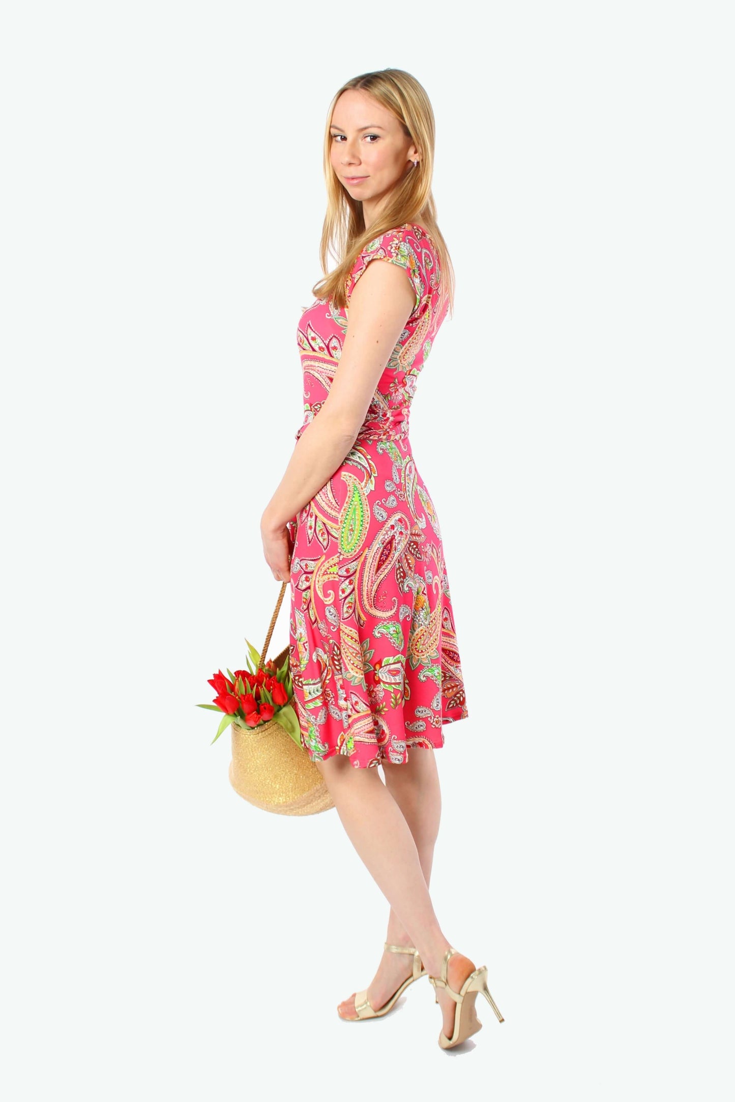 Seitenansicht einer blonden Frau im pinken knielangen Sommerkleid mit Paisley-Muster und Bindegürtel, sie trägt goldene Sandaletten und hält eine Tasche mit Blumen.