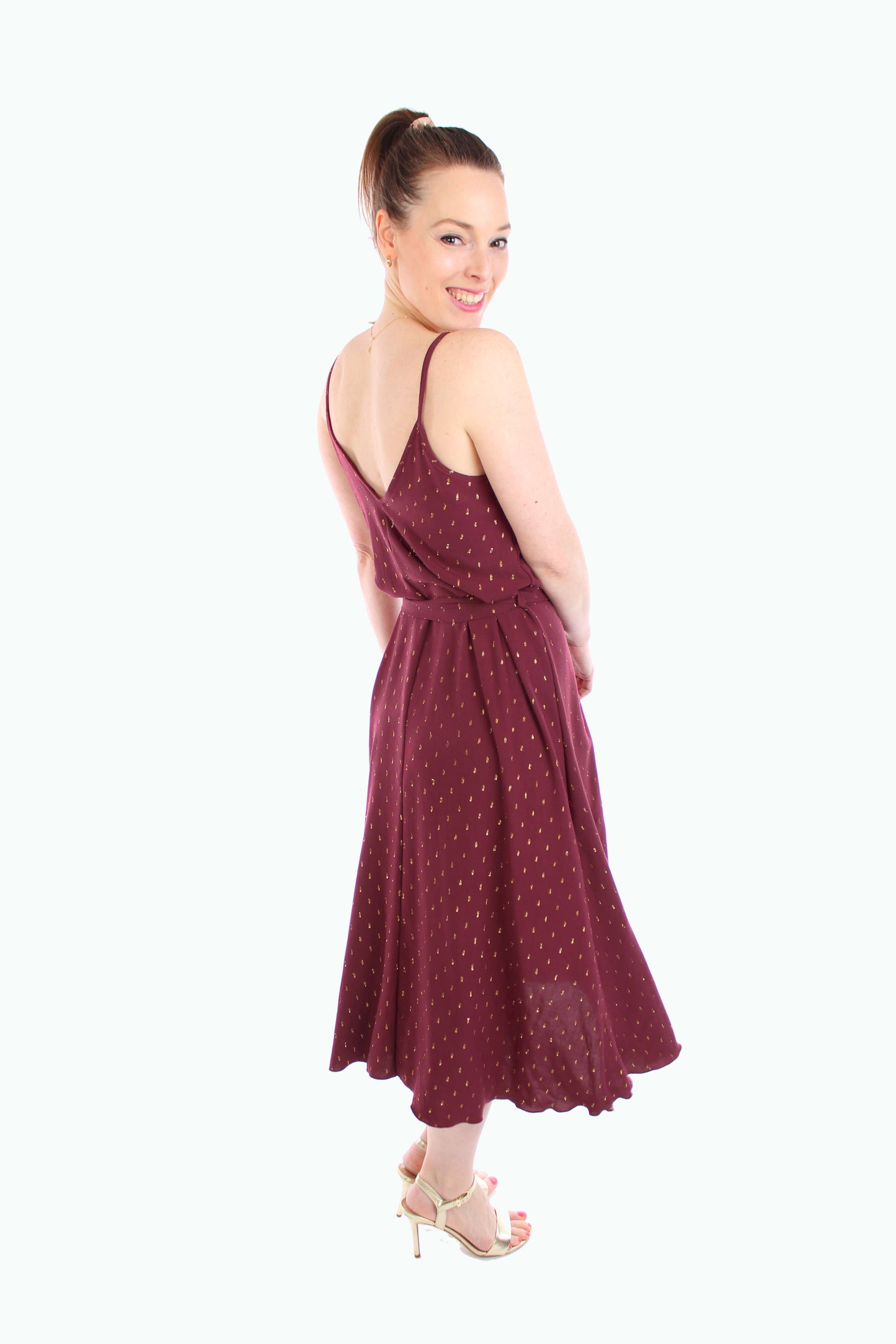 Frau trägt ein bordeauxfarbenes Midikleid mit goldenen Details, Spaghettiträgern und Bindegürtel. Elegantes und luftiges Design, fair produziert in Berlin.