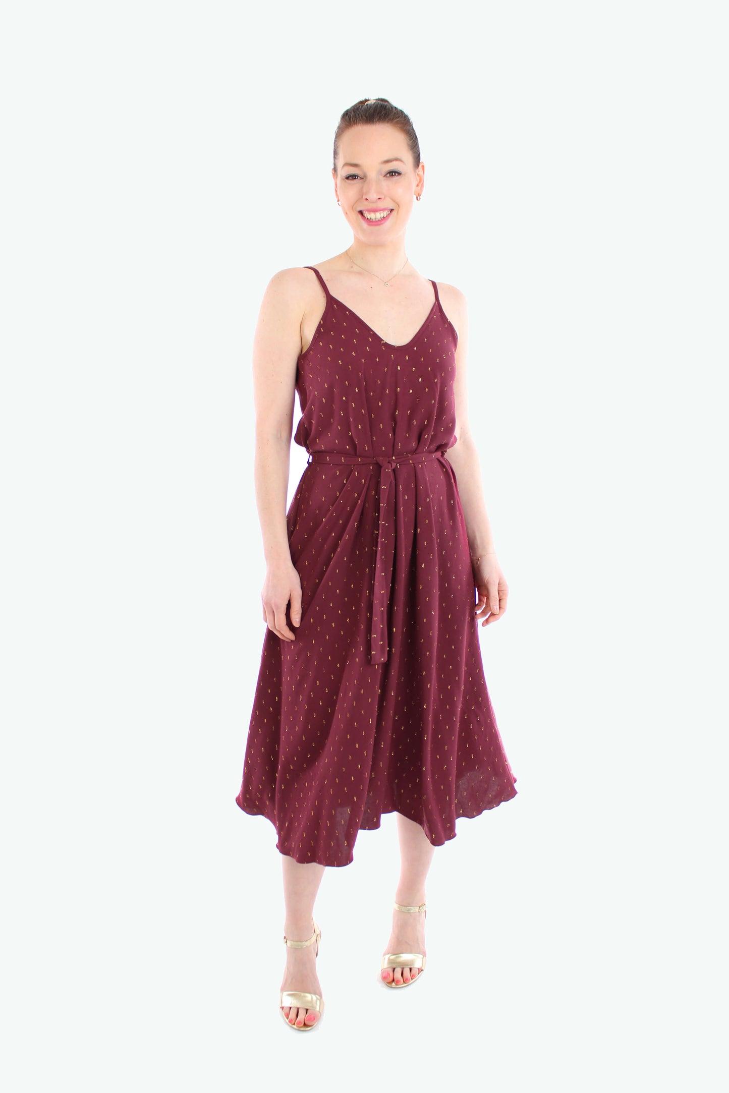 Frau trägt ein elegantes Midikleid 2025 mit Spaghettiträgern in Bordeaux mit goldenen Details. Das luftige Sommerkleid hat einen Bindegürtel in der Taille und fällt fließend bis zur Wade.