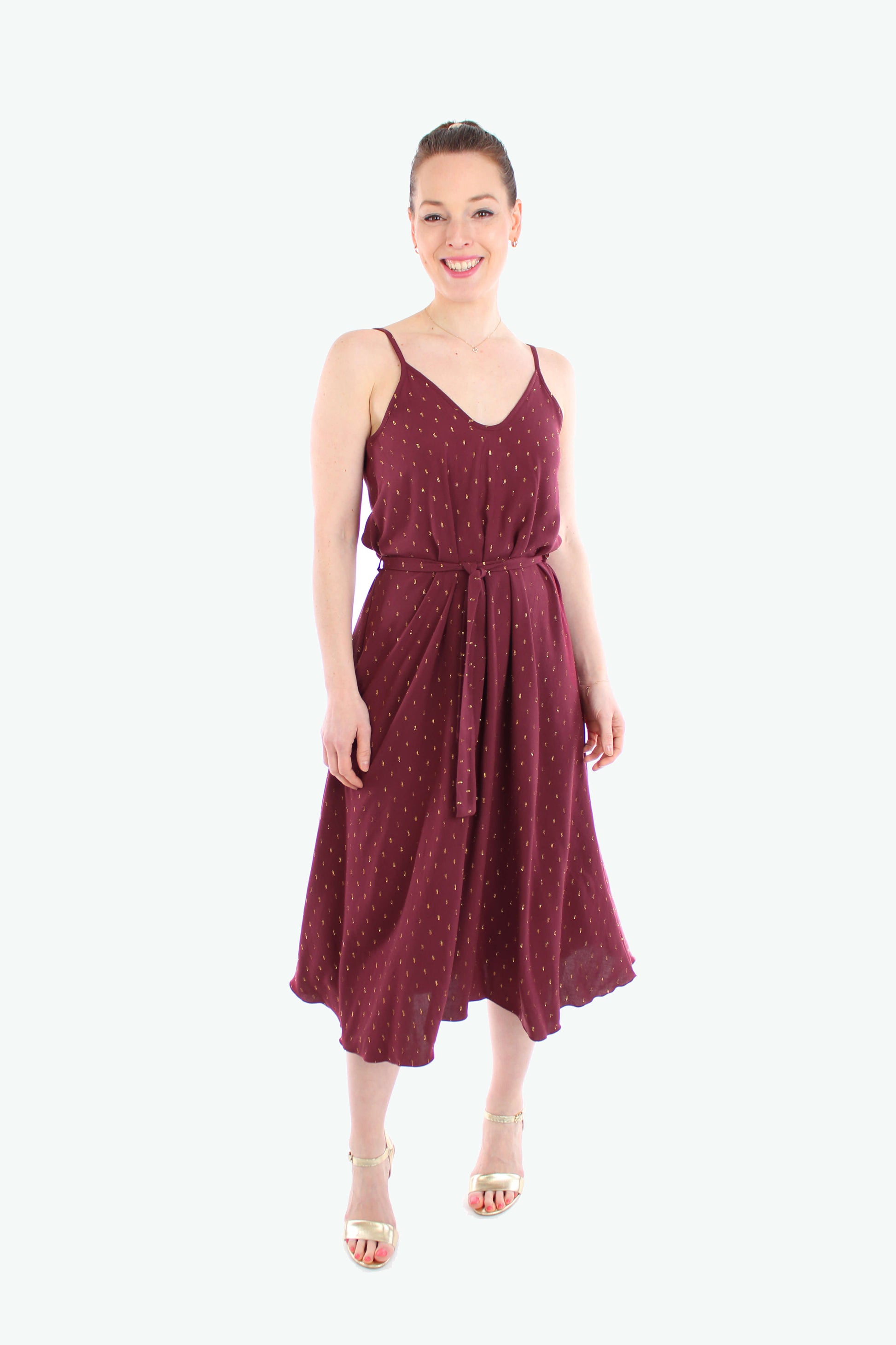 Frau trägt ein elegantes Midikleid 2025 mit Spaghettiträgern in Bordeaux mit goldenen Details. Das luftige Sommerkleid hat einen Bindegürtel in der Taille und fällt fließend bis zur Wade.