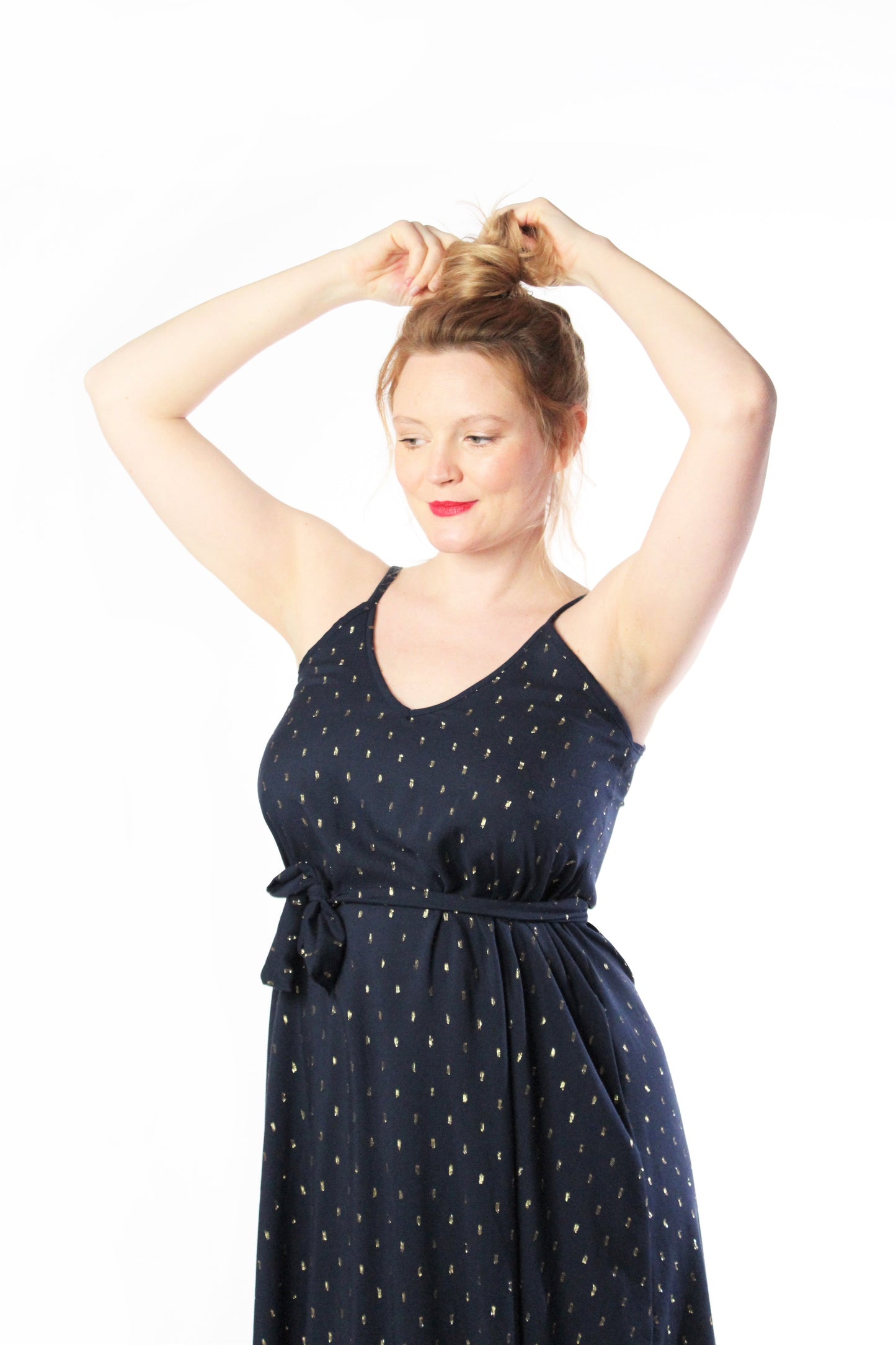 Lächelnde Frau, die sich die Haare hochsteckt. Sie trägt ein dunkelblaues Sommerkleid mit Spaghettiträgern. Das Kleid hat kleine, goldene Punkte und einen schmalen Gürtel.